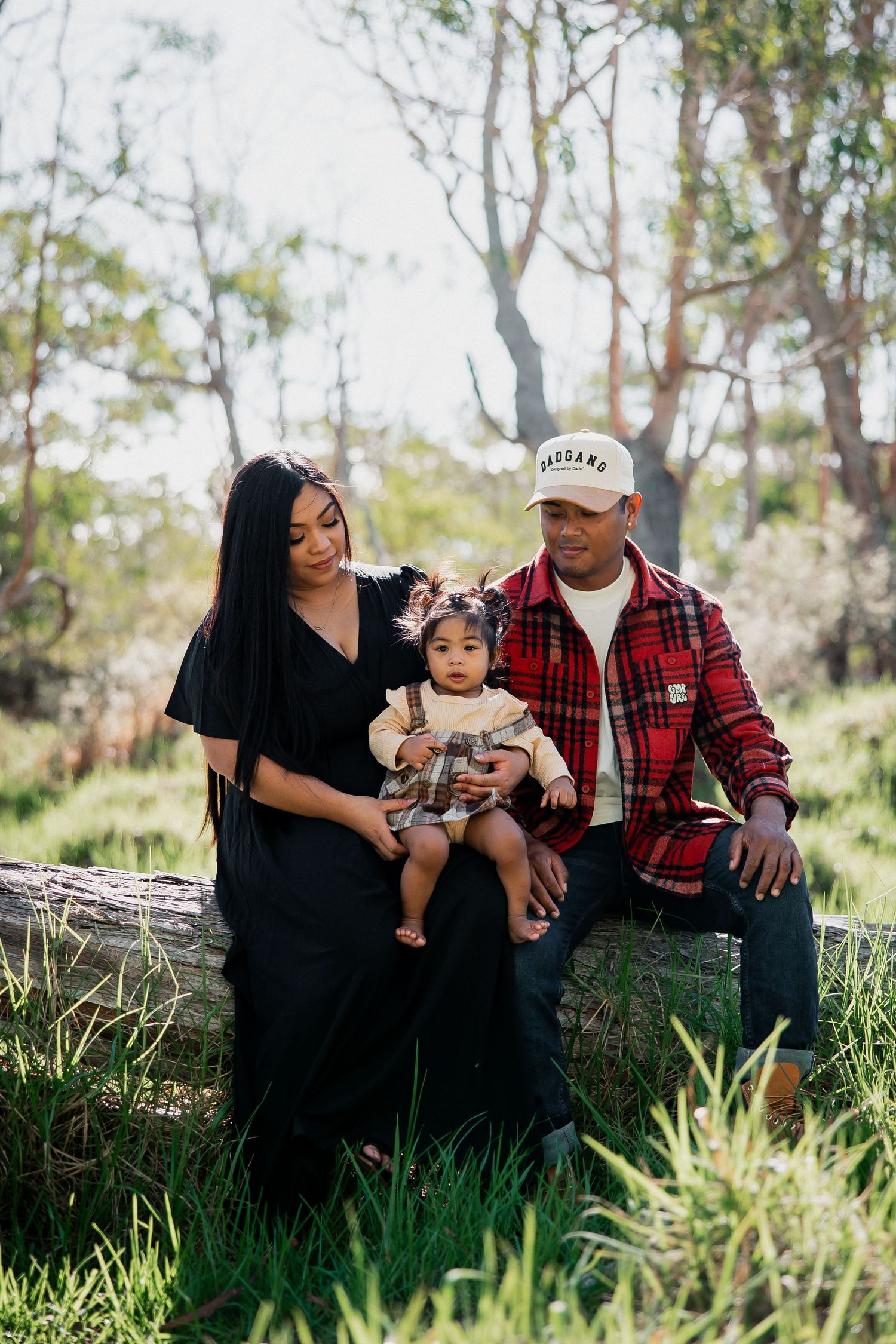 Family of three sitting on a log in a lush green forest, parents in black and plaid outfits with their baby girl.