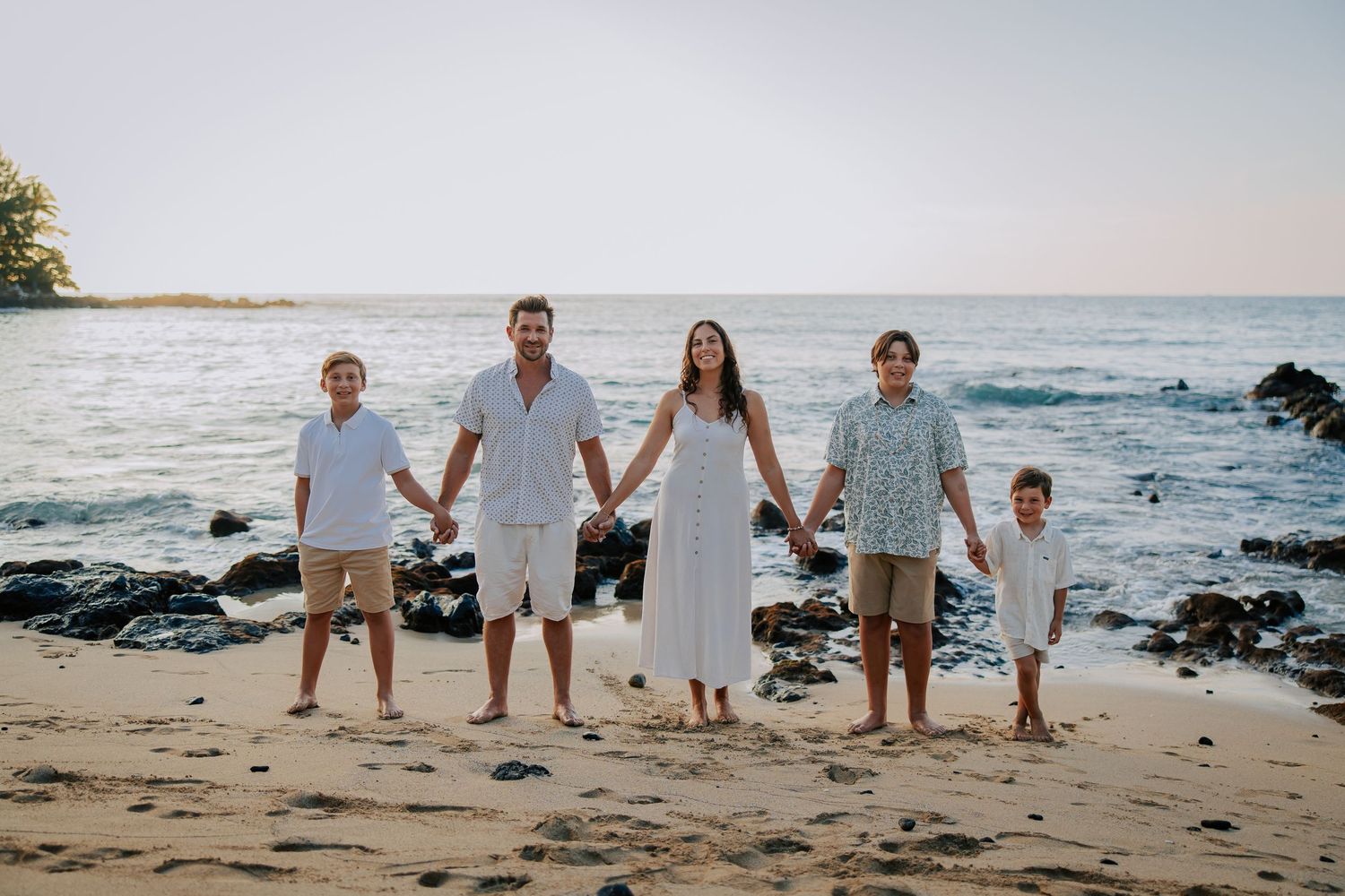 Family of five holding hands on a sandy beach at sunset, wearing white and light blue outfits.
