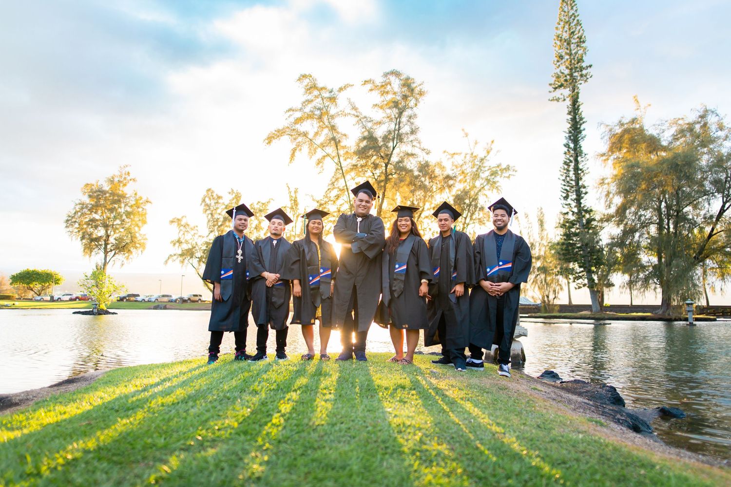 Group of graduates in black caps and gowns posing outdoors by a lake at sunset on graduation day.