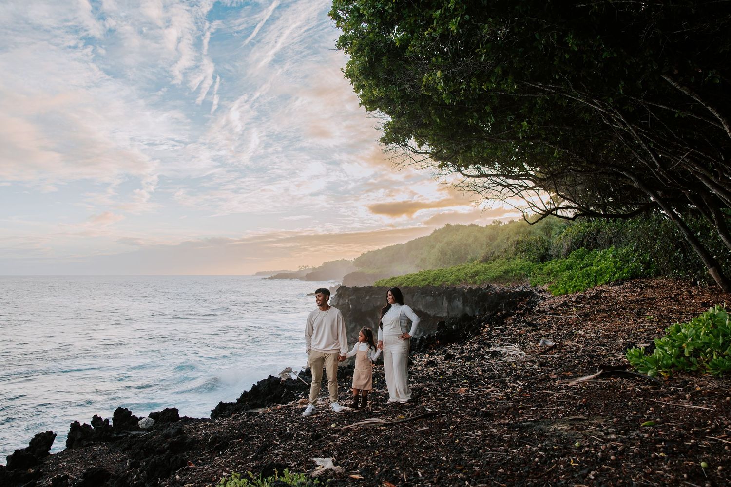 Family in white outfits standing on rocky Hawaiian coastline at sunset with lush green cliffs and ocean views.
