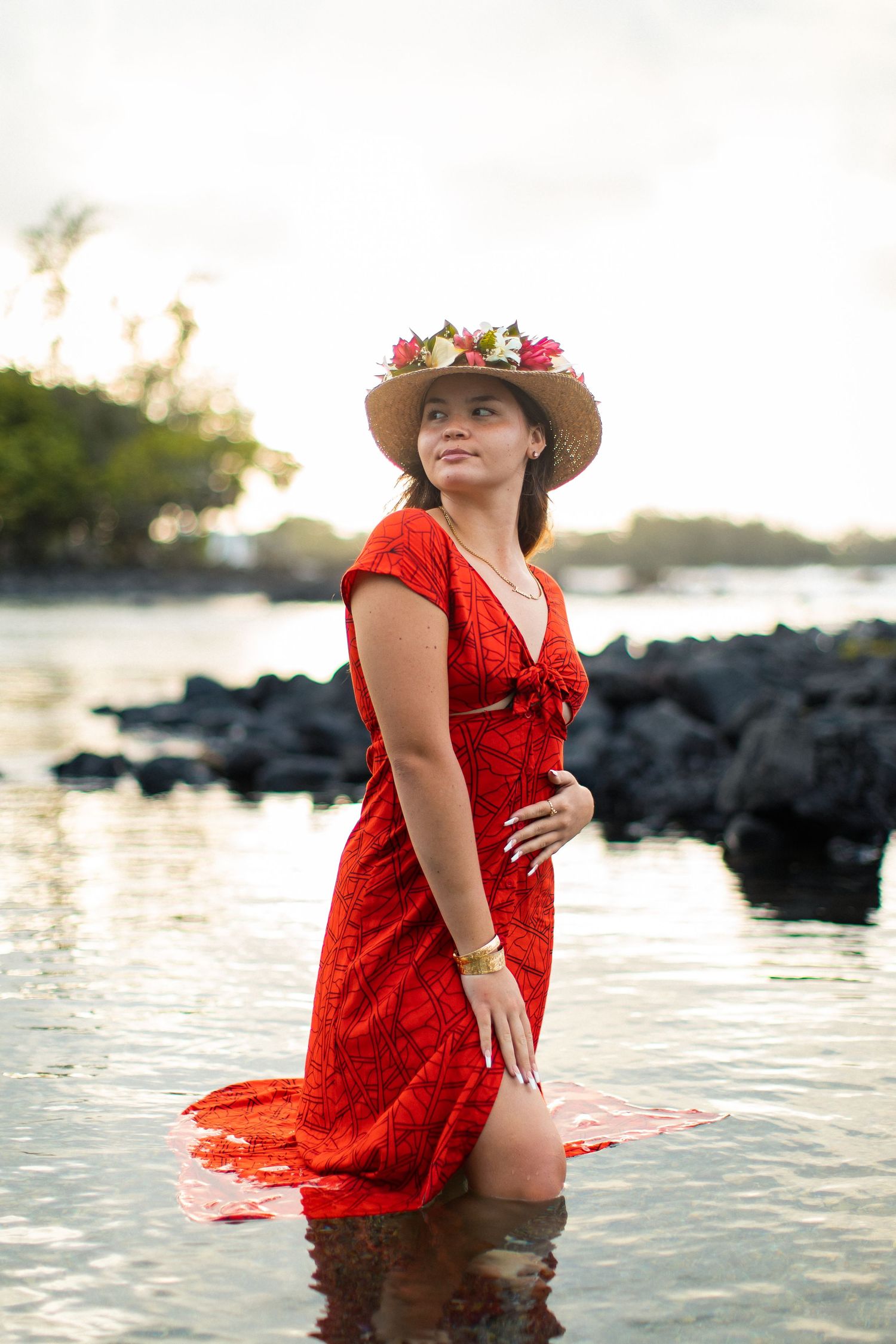 Woman in red dress and floral hat standing in shallow water near rocky shore at sunset.