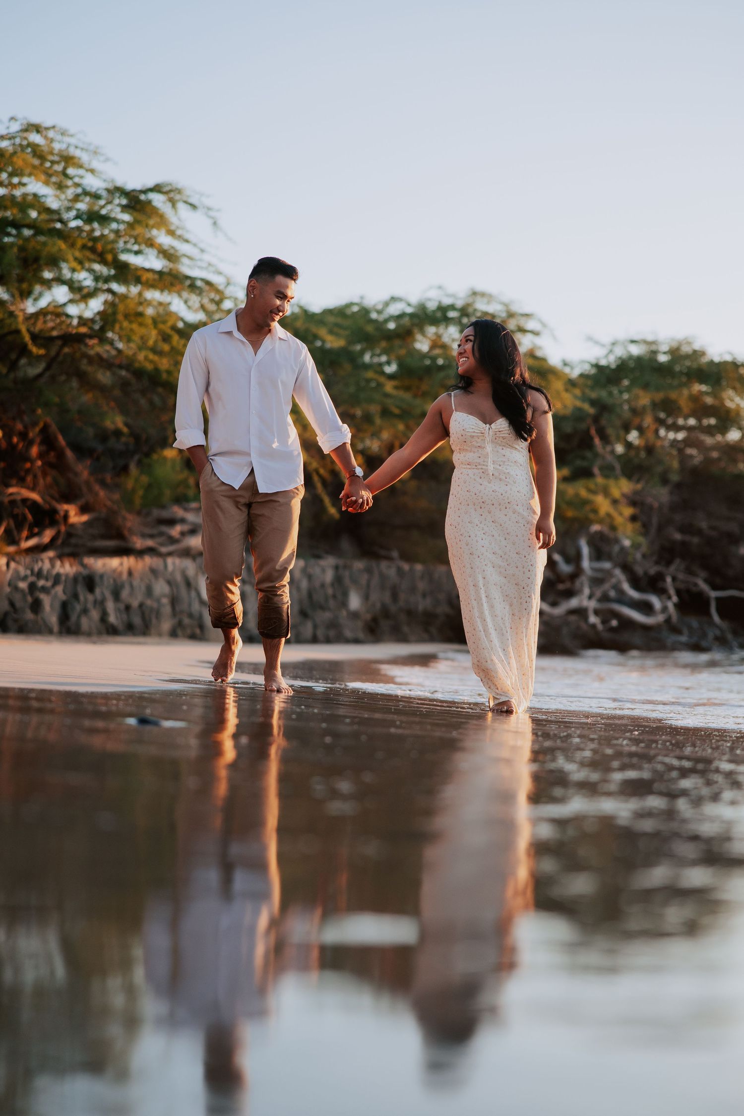 Couple holding hands walking on a reflective beach at sunset, woman in white gown, man in white shirt.