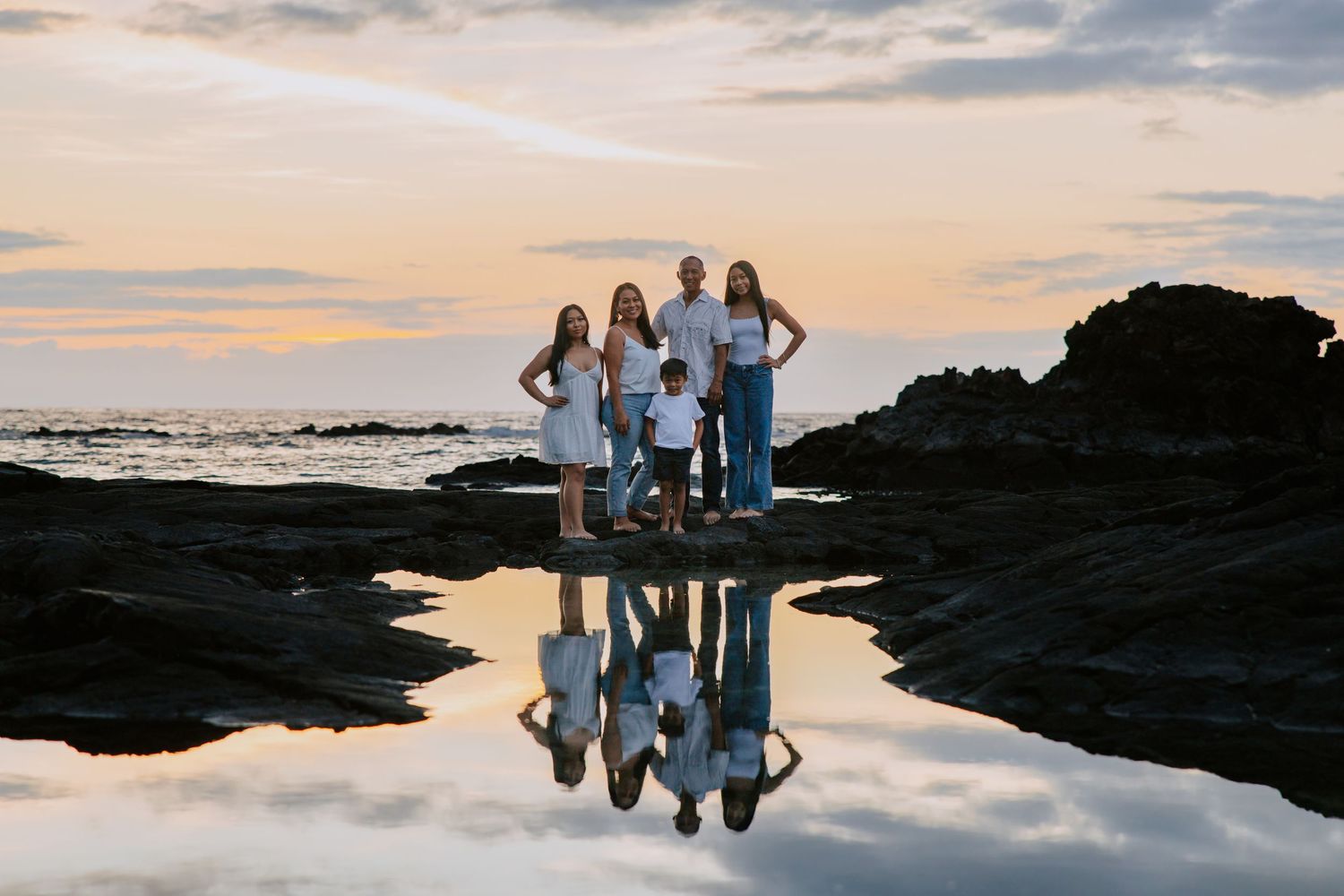 Family of five poses on rocky shore at sunset, their reflection mirrored in a tide pool with ocean waves behind them.