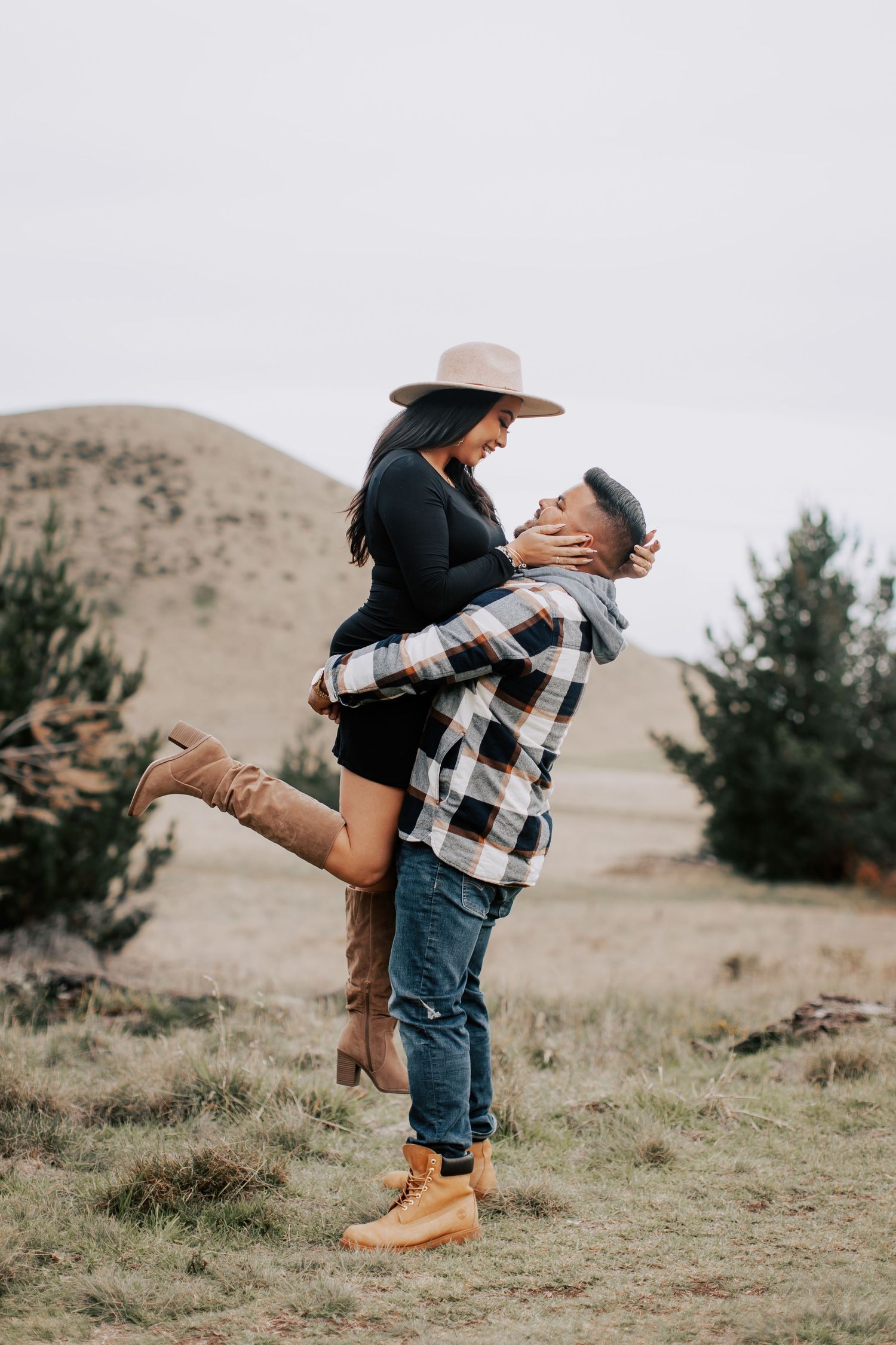A man lifts a woman in a joyful embrace outdoors, both wearing hats and boots in a scenic rural landscape.