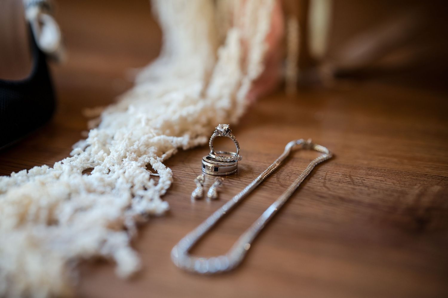 Close-up of wedding rings and diamond necklace on wooden floor beside a white lace bridal scarf.
