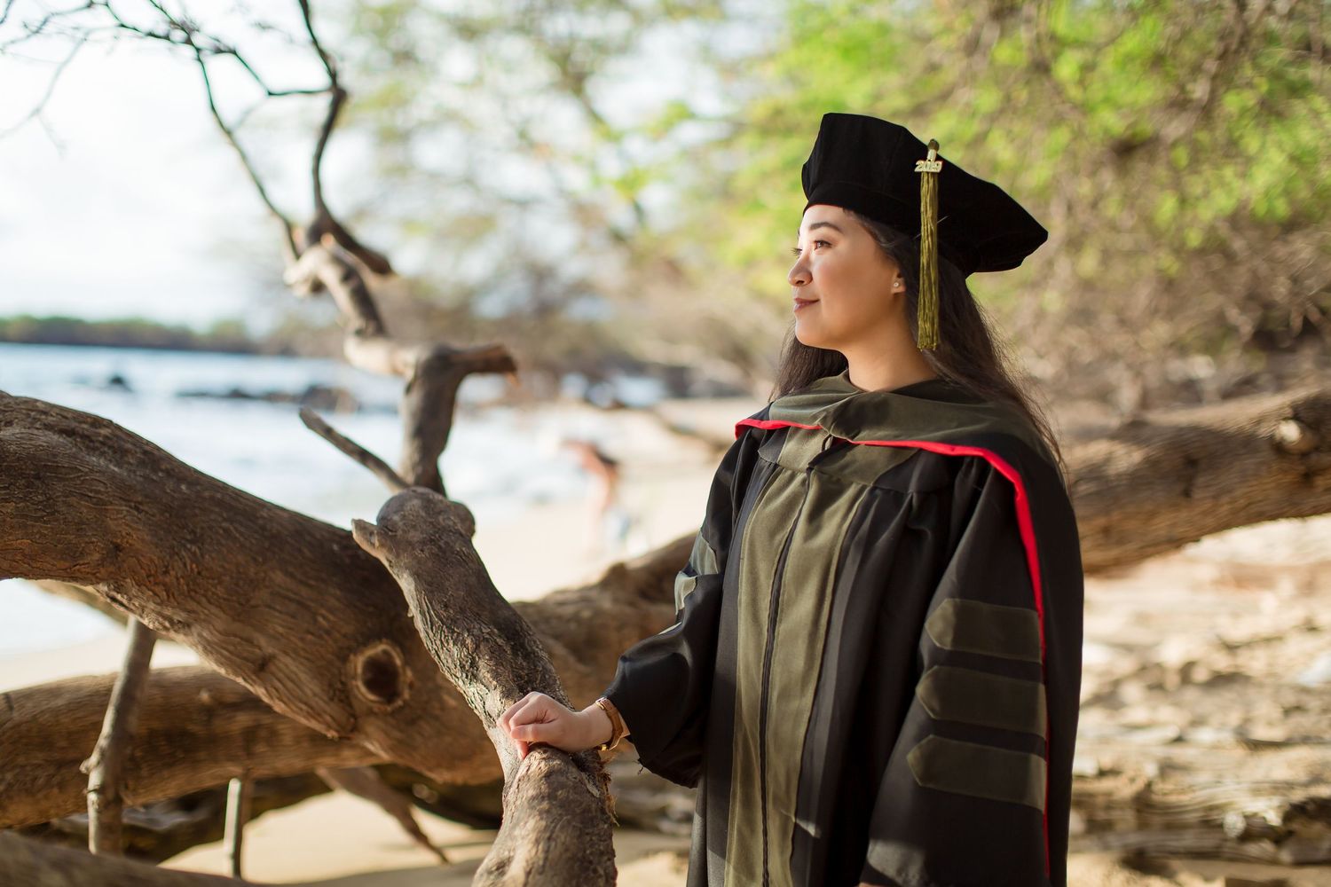 Woman in graduation cap and gown posing outdoors near driftwood on a tropical beach.