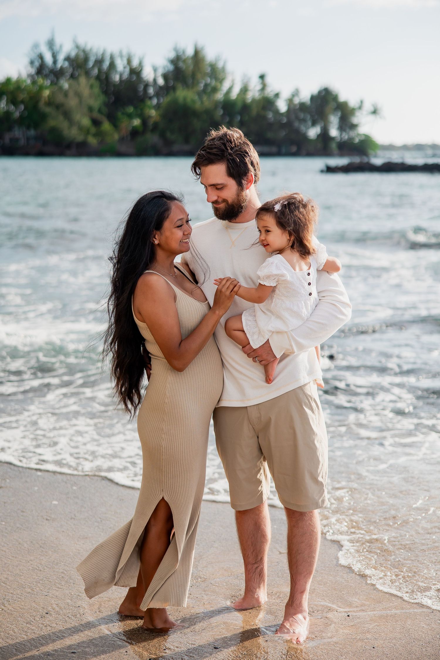 A happy family of three poses barefoot on a tropical beach, with parents holding their young child near the ocean.