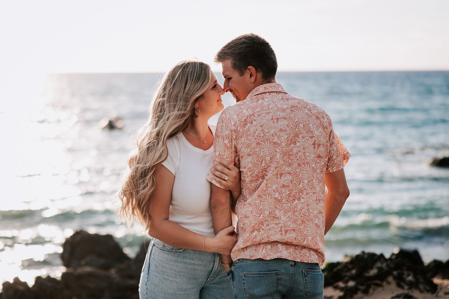 Couple sharing a romantic moment by the ocean, woman with long blonde hair in white top and man in floral shirt.