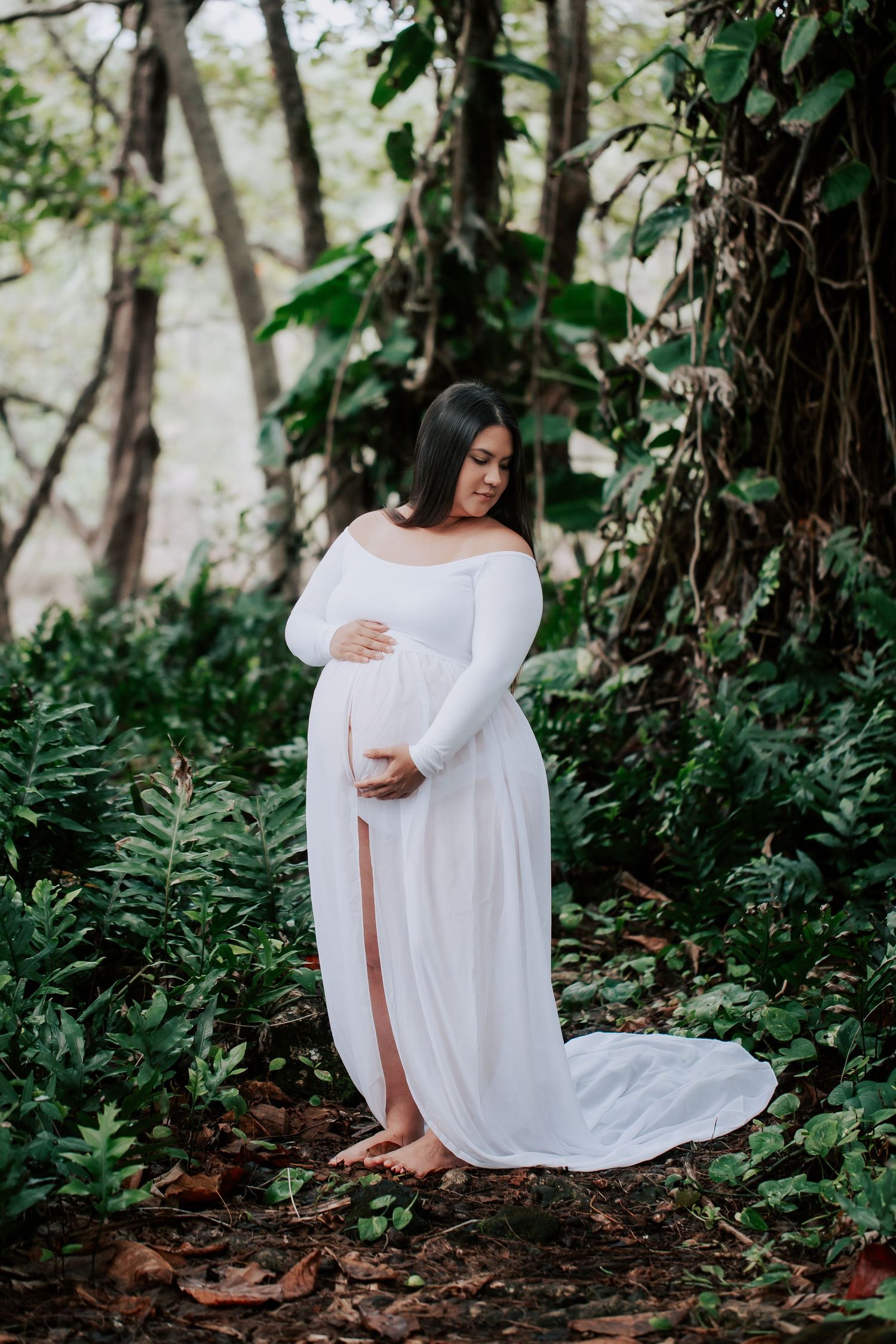 Pregnant woman in white off-shoulder gown cradling belly outdoors in lush green forest setting.