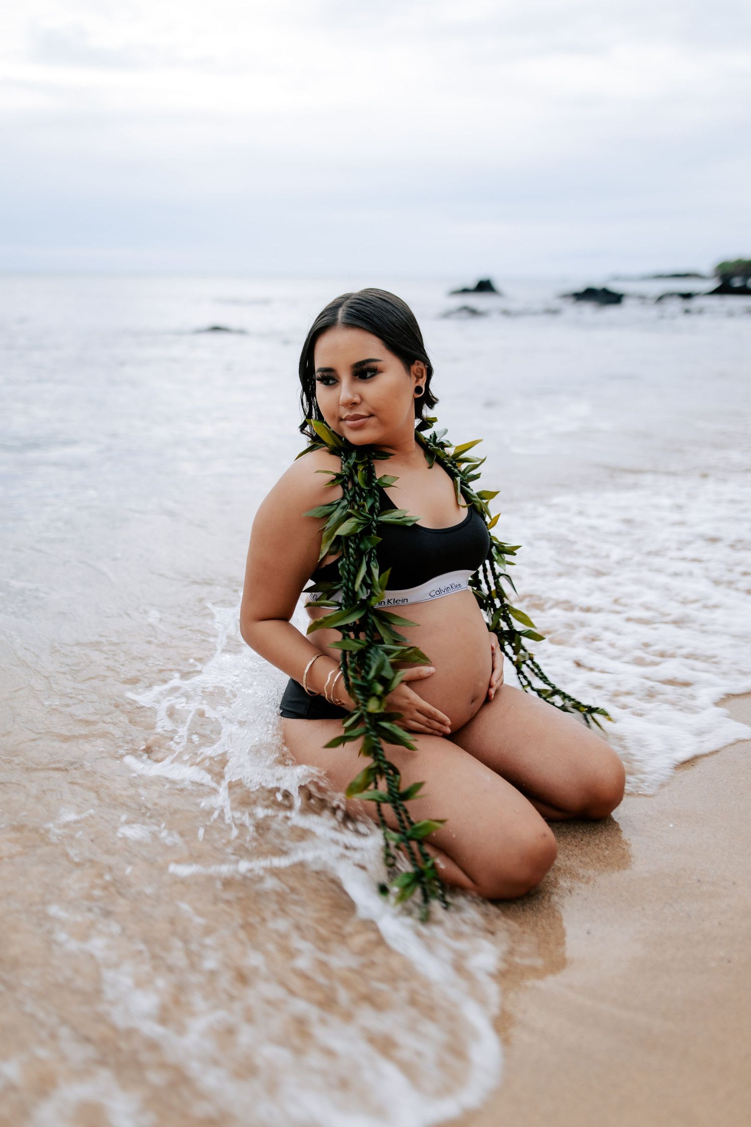 Pregnant woman in black bikini with green lei sits on sandy beach shore with ocean waves, maternity photo shoot.