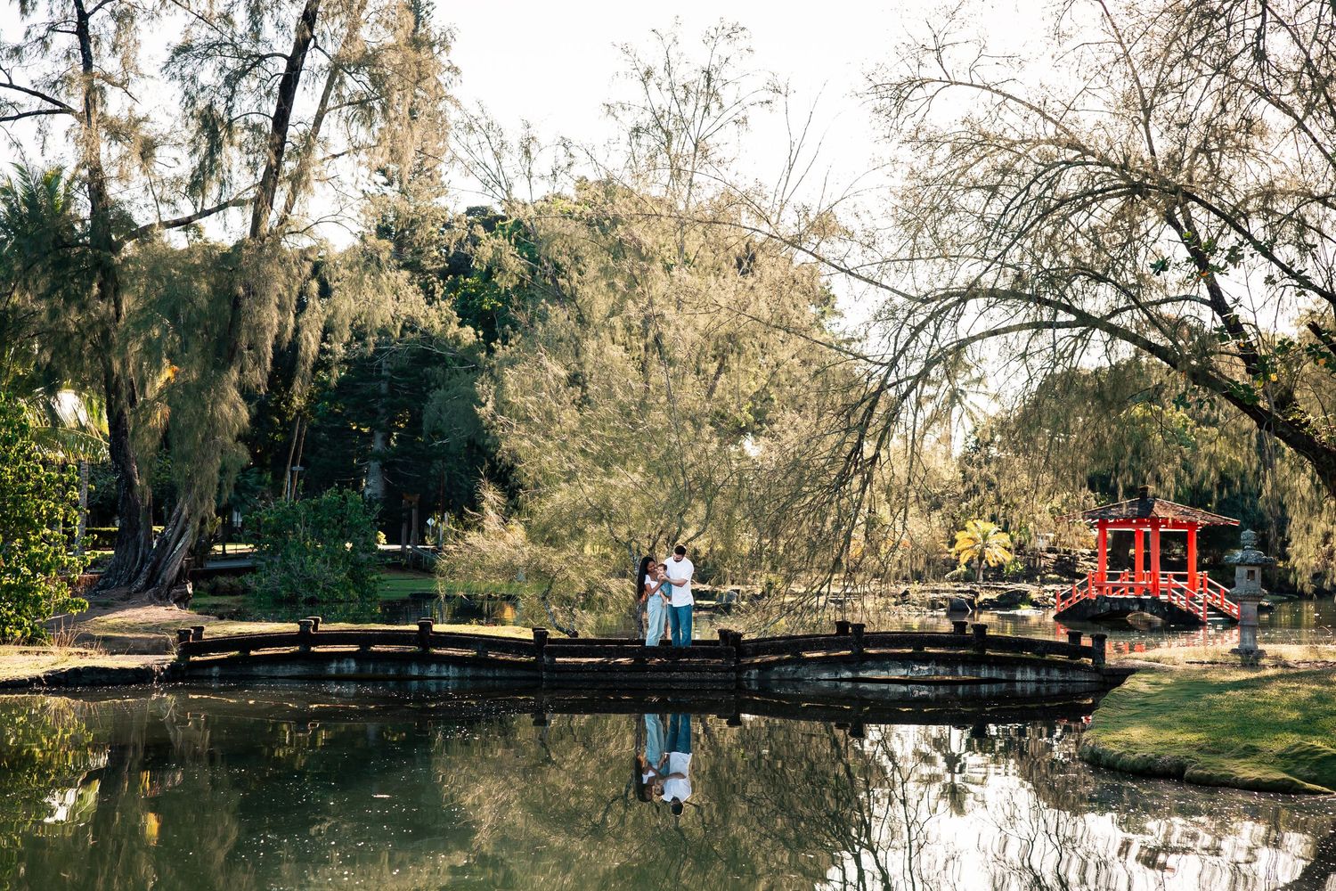 Couple embracing on a wooden bridge over a tranquil pond in a lush Japanese garden with a red torii gate.