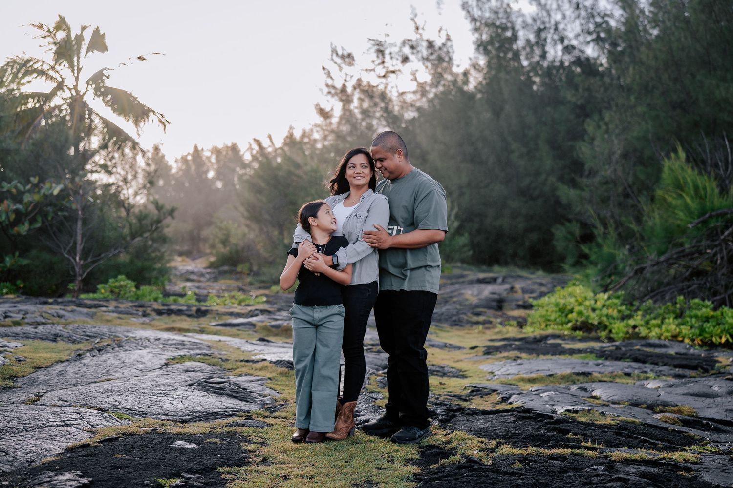 Family of three posing on rocky Hawaiian shoreline at golden hour, surrounded by lush tropical greenery.