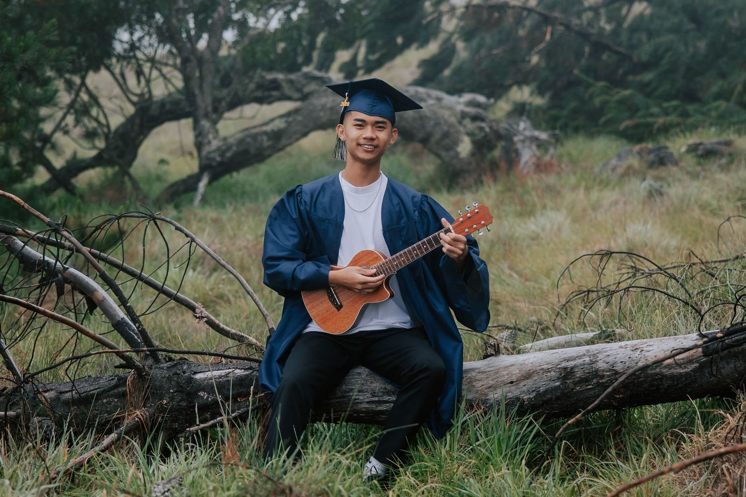 Graduate in cap and gown plays ukulele outdoors, sitting on a fallen log in a grassy field.