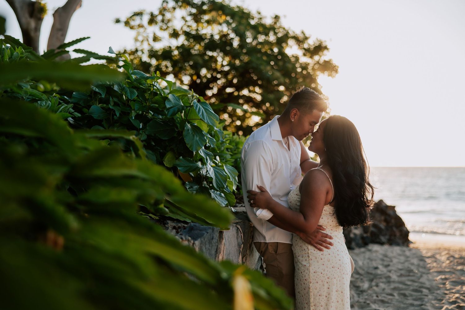 Couple sharing a romantic sunset kiss on a tropical beach surrounded by lush green foliage and ocean waves.
