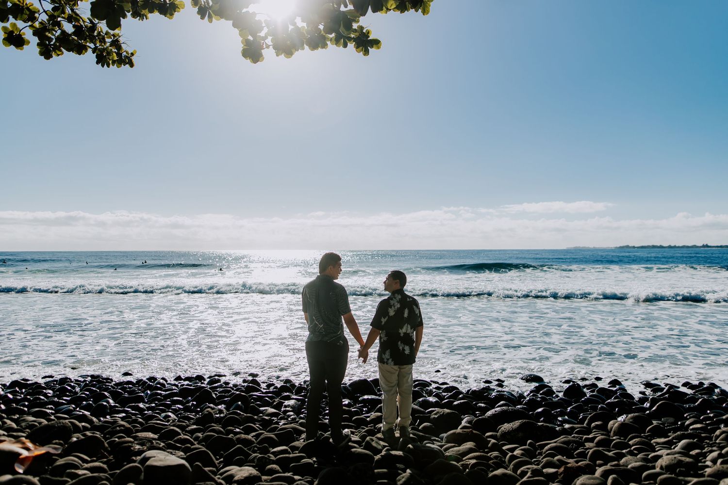 Elderly couple holding hands on rocky Hawaiian beach, standing before sparkling ocean waves under bright sunny sky.