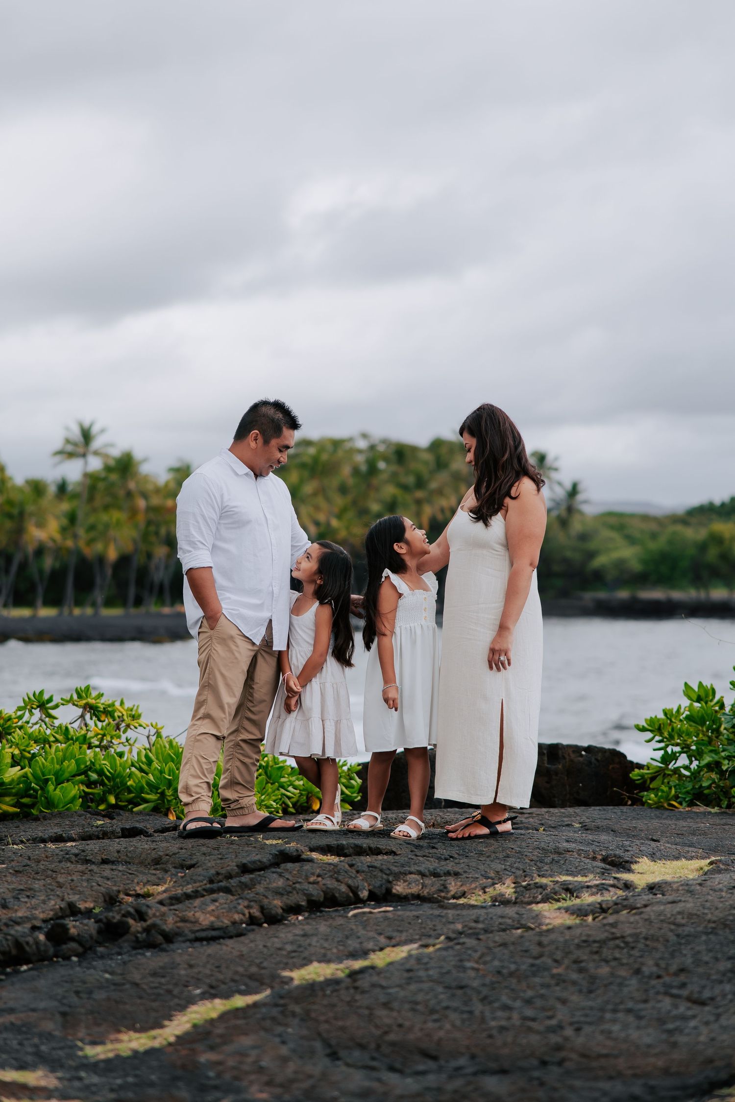 Family of four in white outfits posing on volcanic rock by the ocean with tropical palm trees in Hawaii.