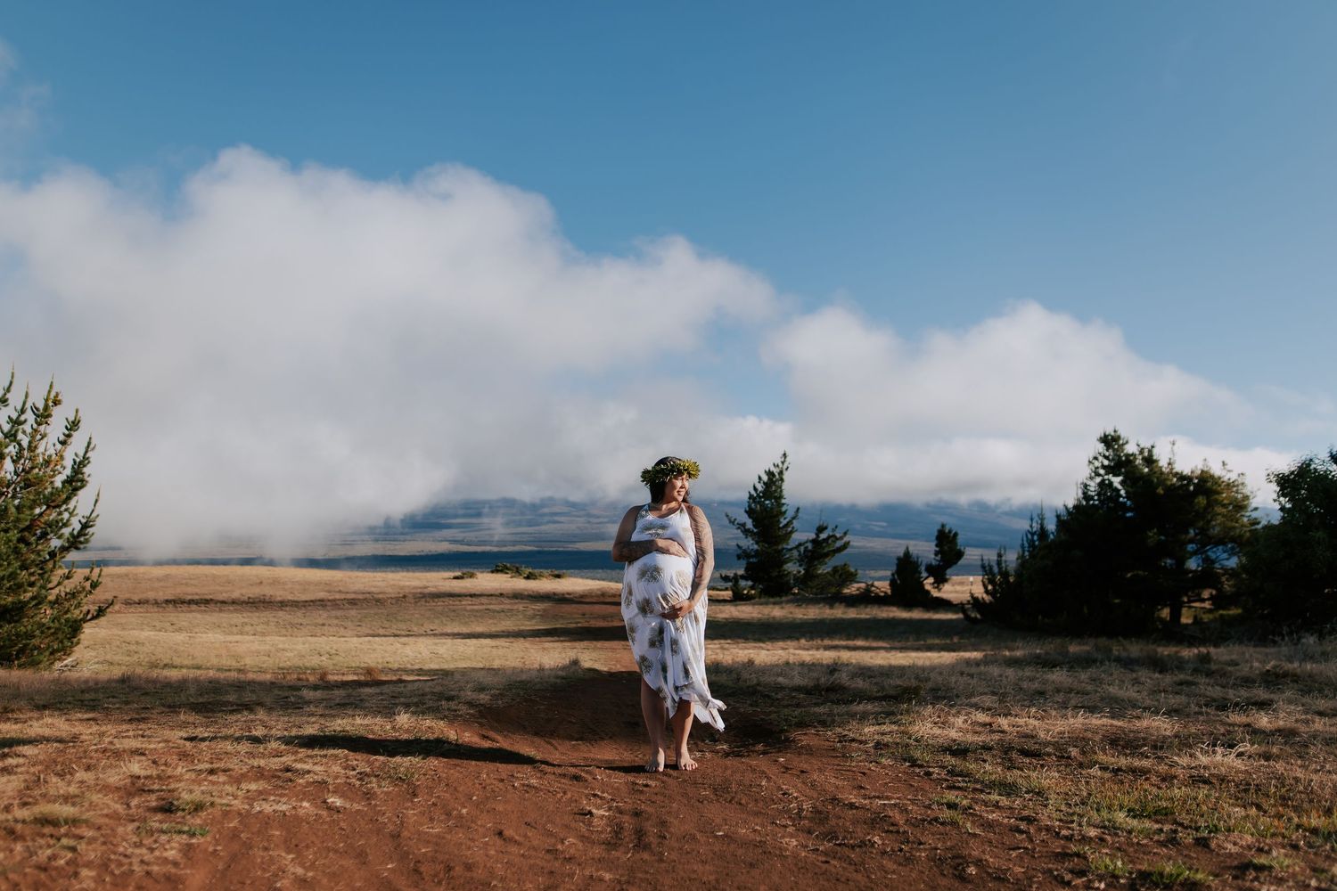 Pregnant woman in floral dress stands on red dirt path with mountain landscape and clouds behind her.