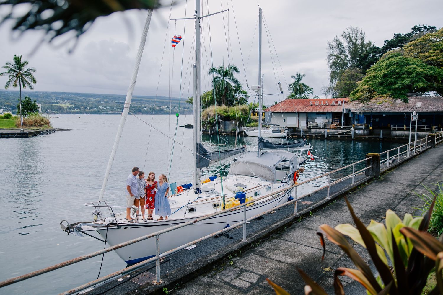 Family of three posing on a white sailboat docked at a tropical marina with palm trees and calm waters.
