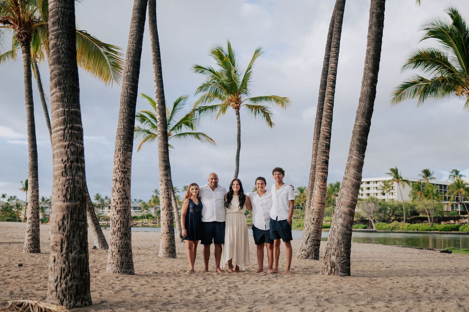 Family of five posing among tall palm trees on a sandy beach with tropical resort in background.