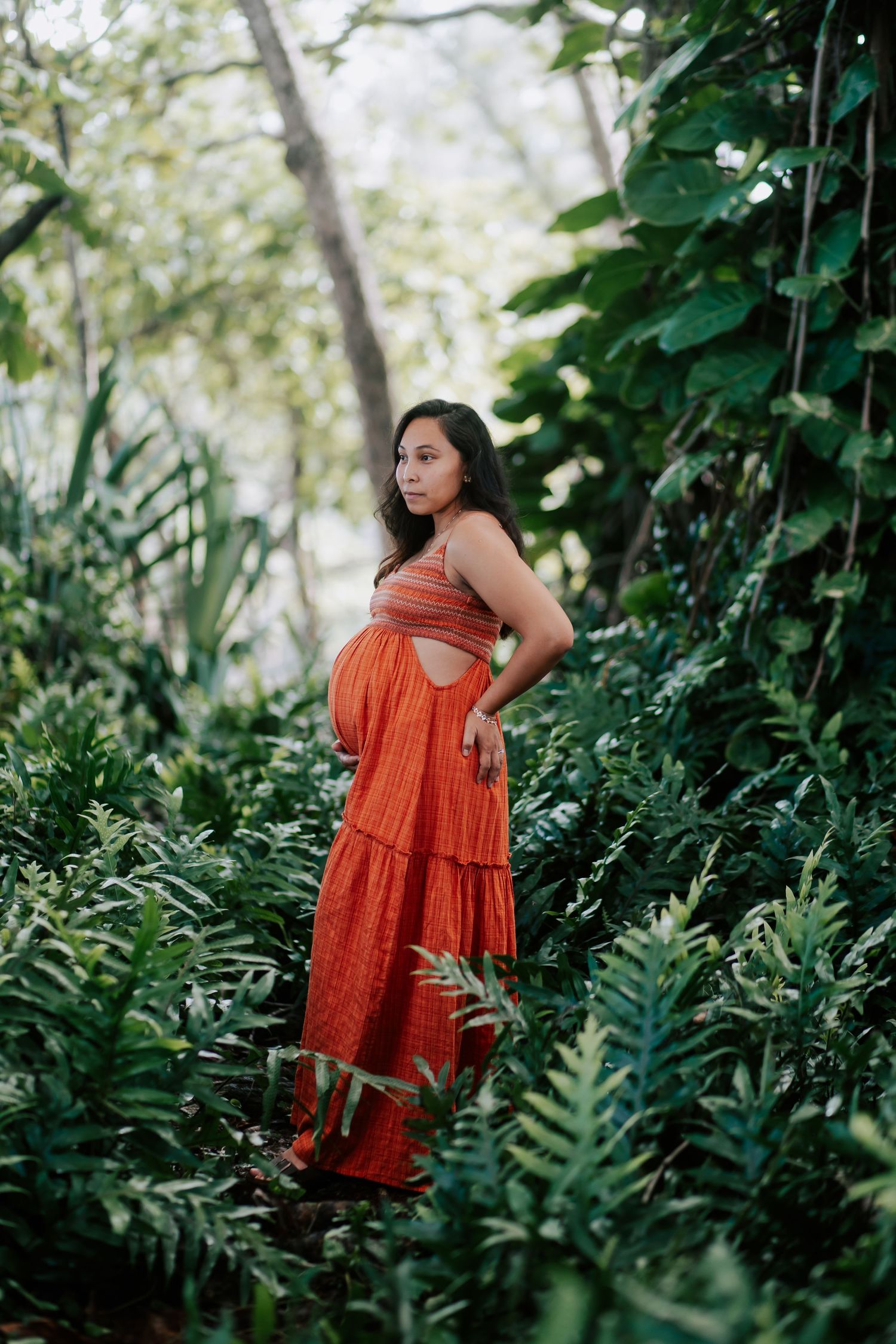 Pregnant woman in orange maxi dress standing among lush tropical greenery during maternity photoshoot.