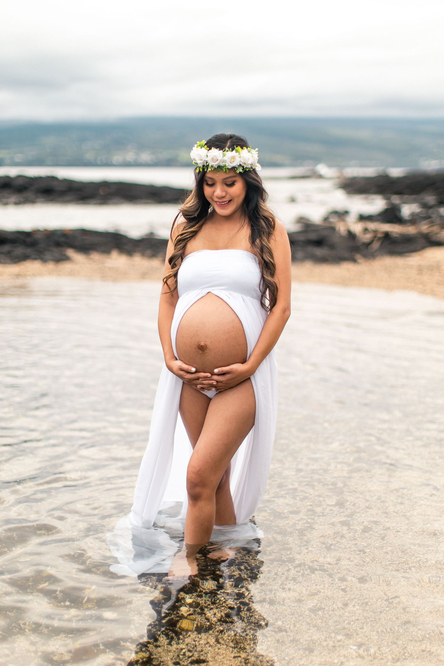 Pregnant woman in white wrap dress and floral crown stands barefoot in ocean water during a maternity photo shoot in Hawaii.