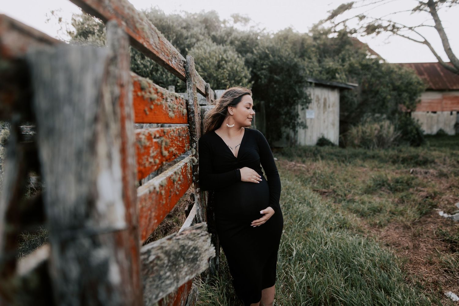 Pregnant woman in black dress leaning against rustic brick wall outdoors, cradling her baby bump in a serene rural setting.