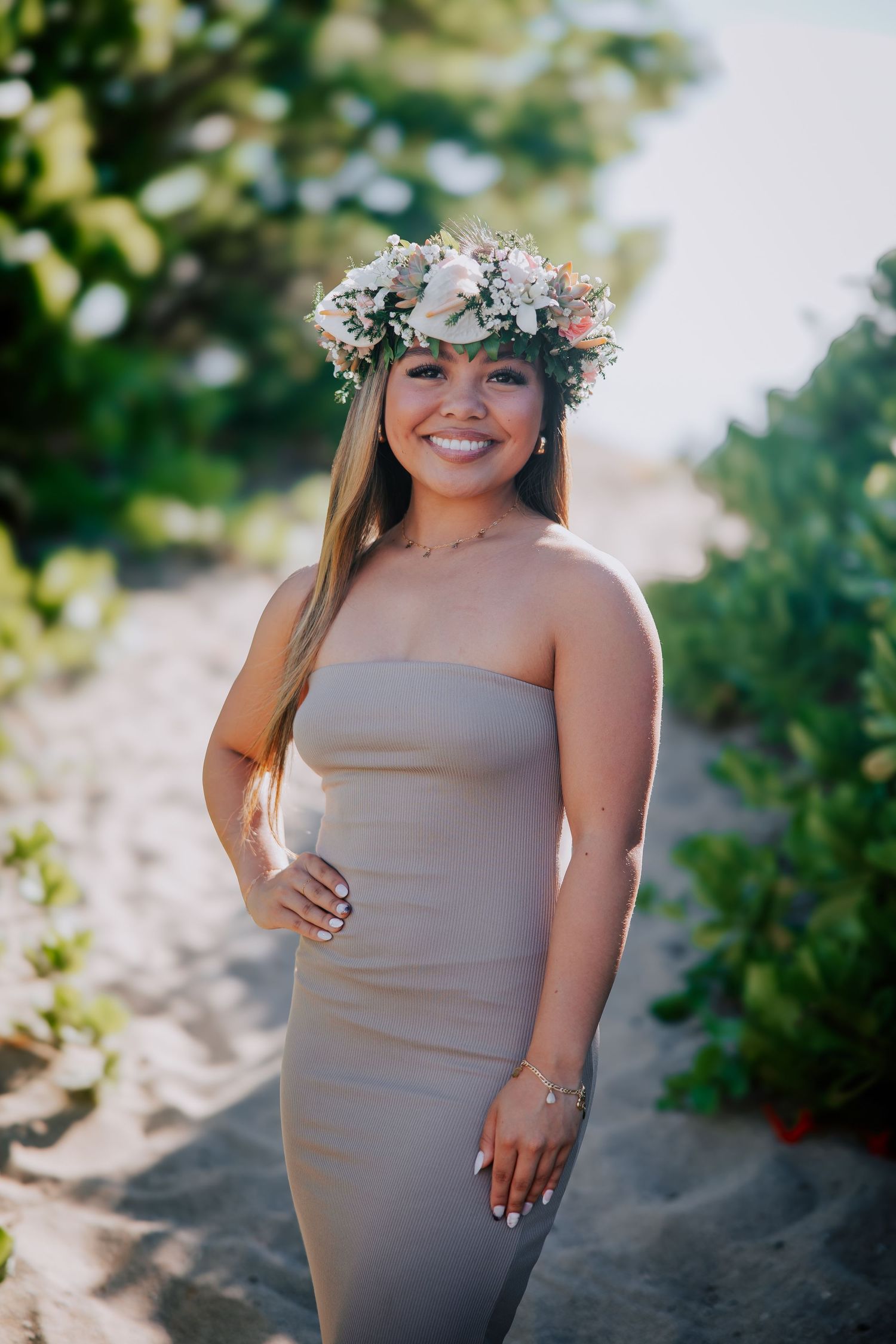 Smiling woman in strapless beige dress wearing floral crown on sandy beach path surrounded by tropical greenery.