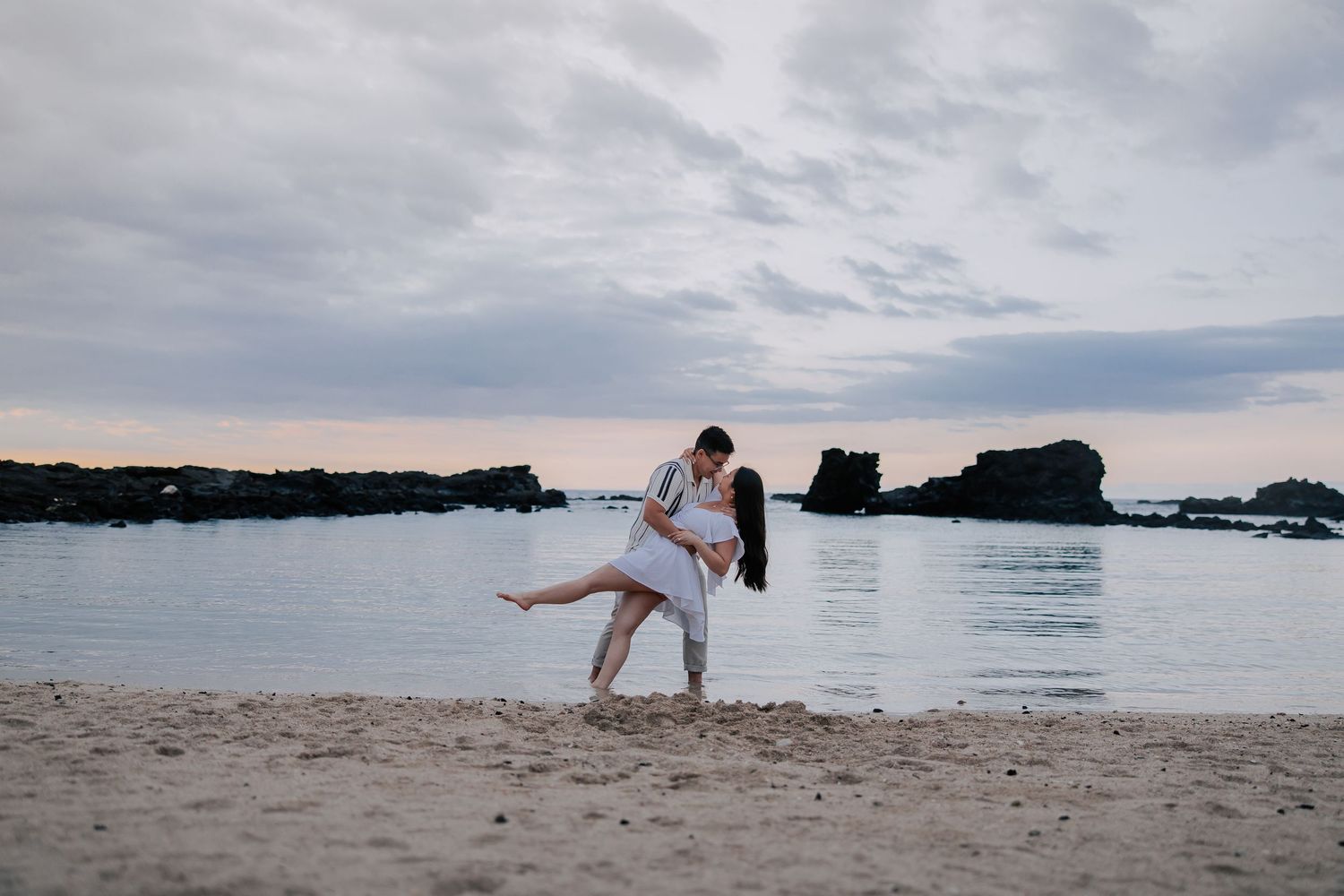Romantic couple embracing on a sandy beach at sunset with rocky ocean backdrop.