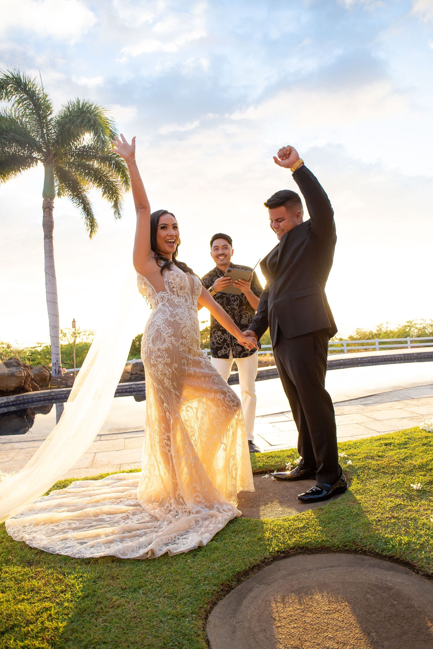 Joyful bride and groom celebrate with raised arms after outdoor wedding ceremony near tropical palm trees at sunset.