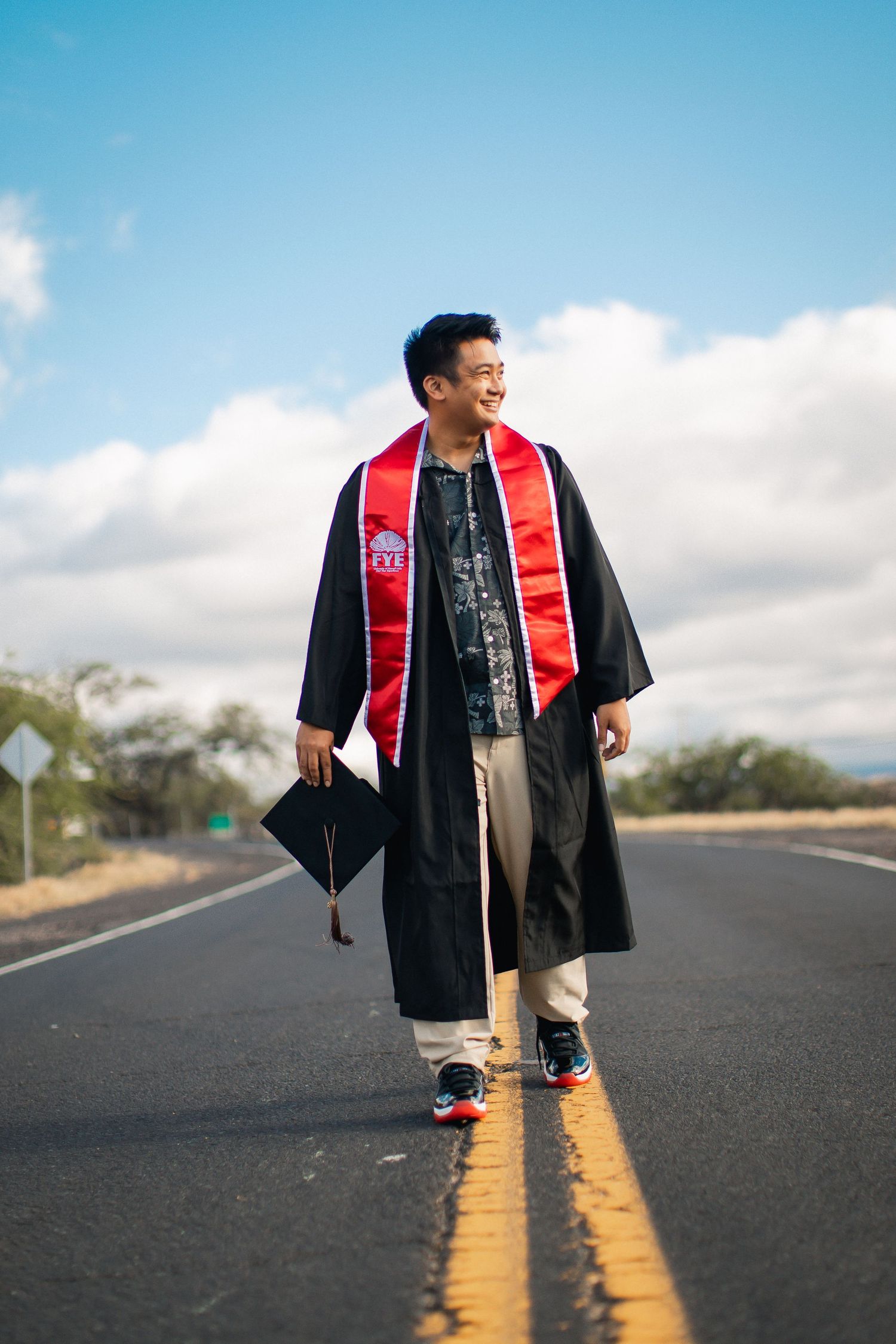 A young man in black graduation gown and red sash walks confidently down an open road holding his cap under a blue sky.
