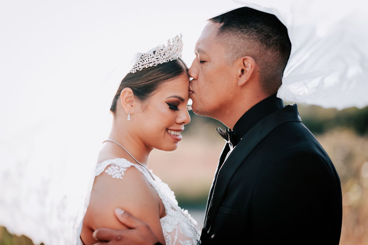A bride wearing a tiara and groom in a black suit share a romantic moment outdoors at their wedding.