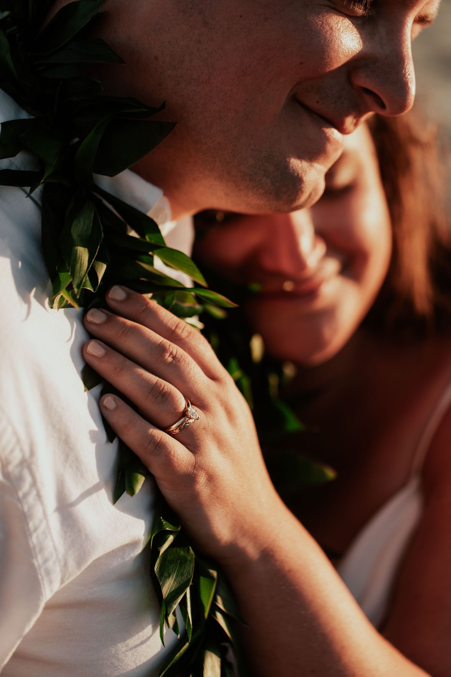 Smiling couple in a romantic close-up, woman wearing an engagement ring and holding green lei garland in warm golden light.