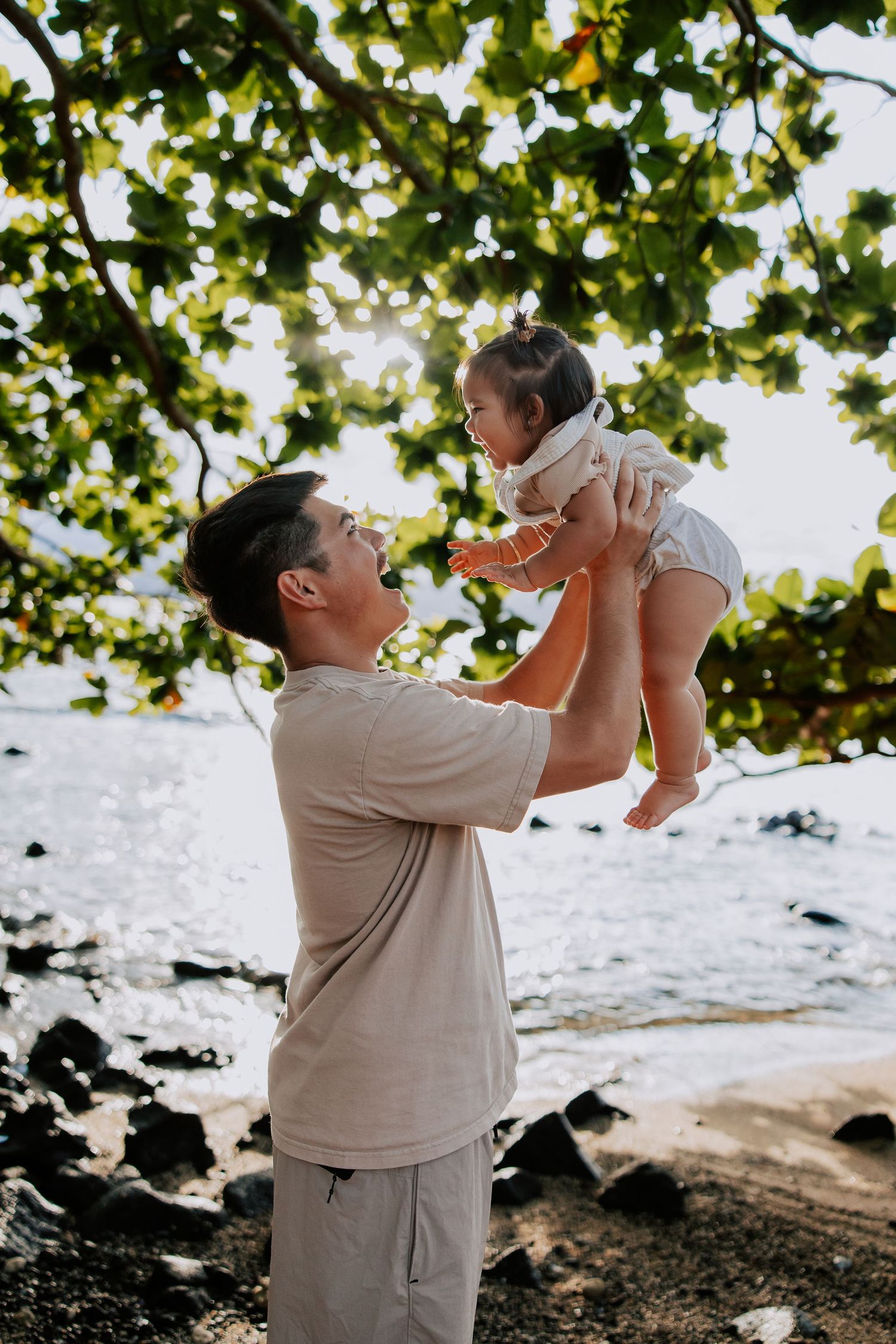 Father joyfully lifts smiling baby in the air on a rocky beach, with sunlight filtering through lush green trees.