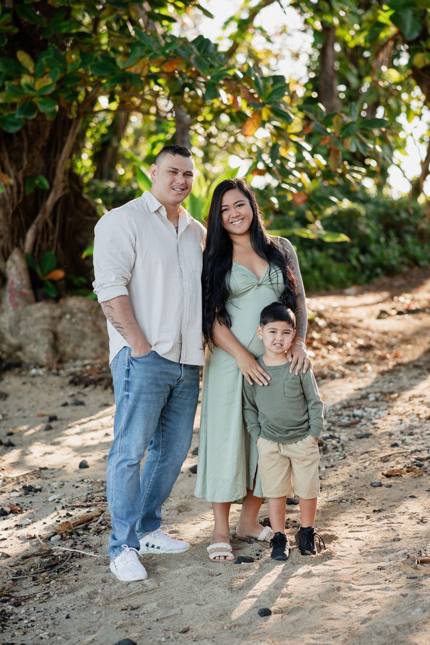 Family of three posing outdoors on sandy path with lush green trees, parents smiling with young son between them.