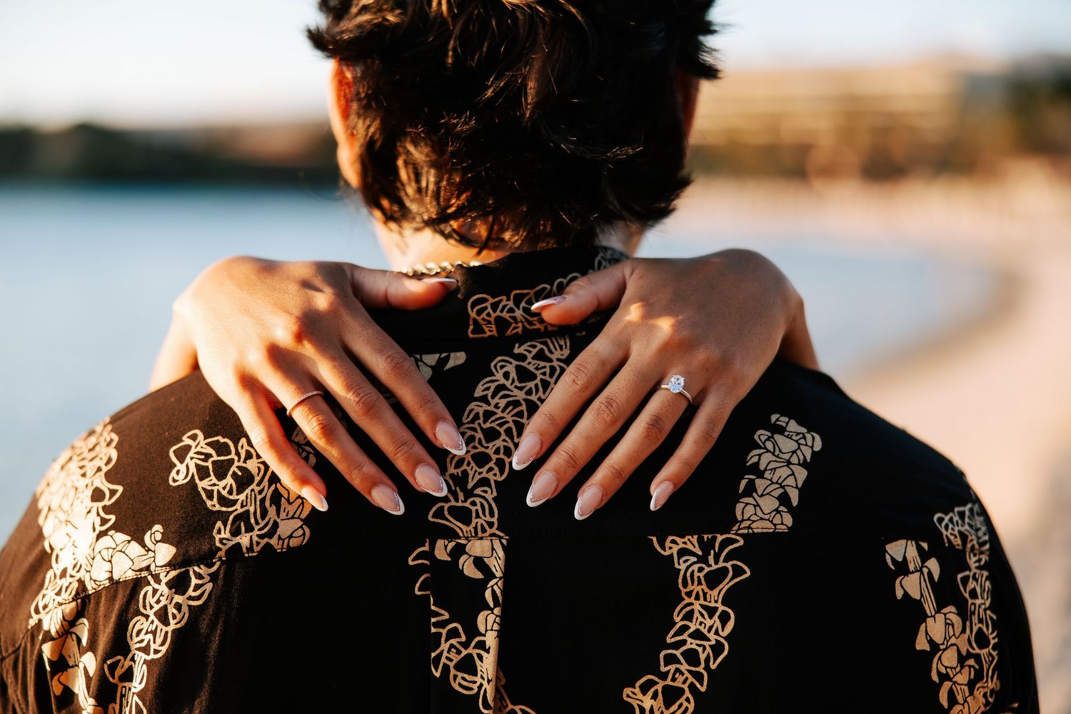 Woman with diamond engagement ring embracing partner wearing ornate black and gold embroidered jacket near waterfront.