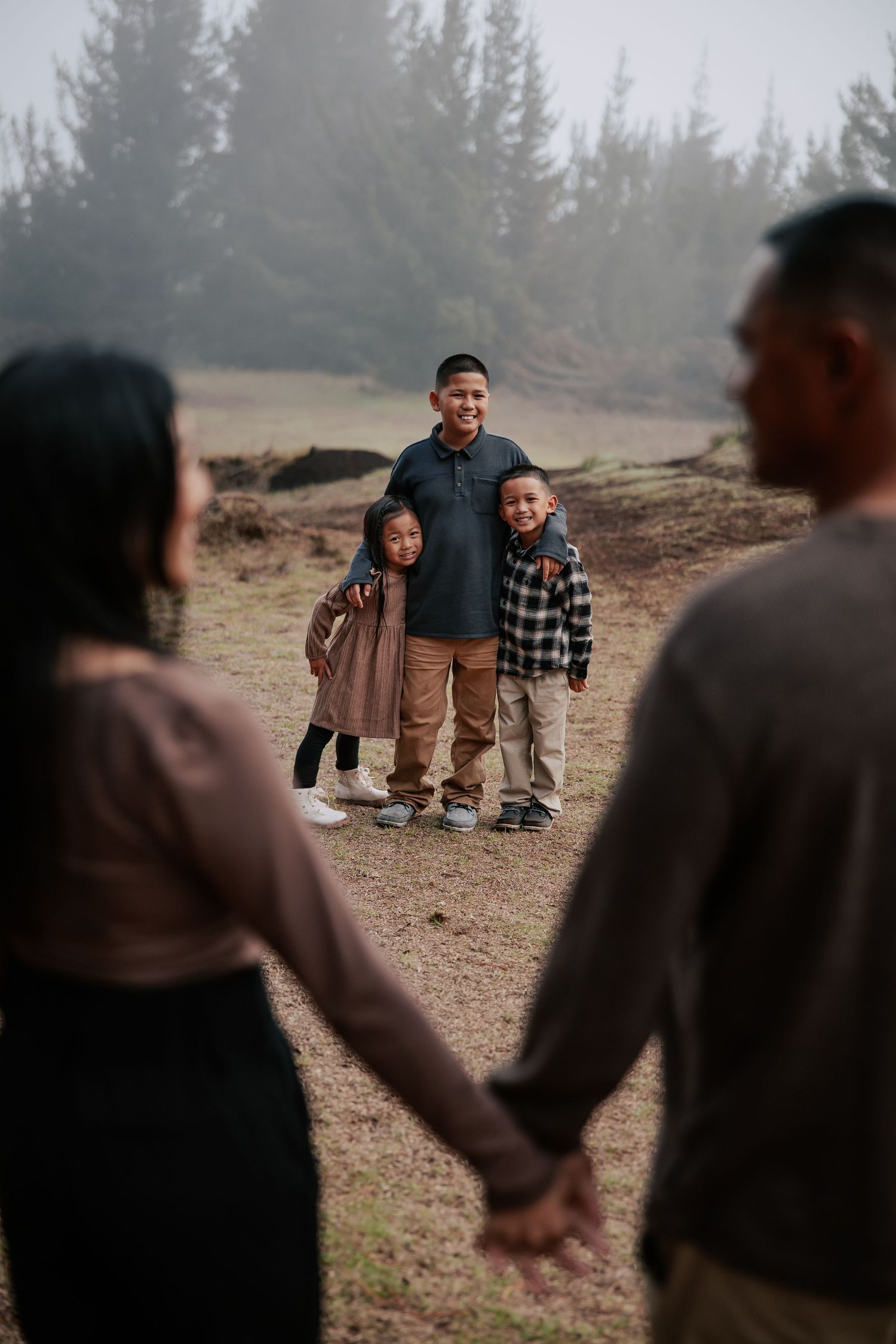 Family of four holding hands outdoors in misty forest, parents in foreground, two children smiling in background.