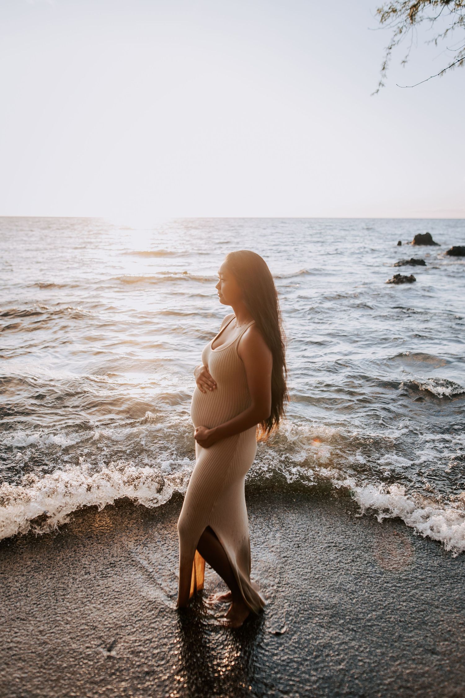 Pregnant woman in beige dress standing at ocean shore during golden sunset, cradling baby bump.