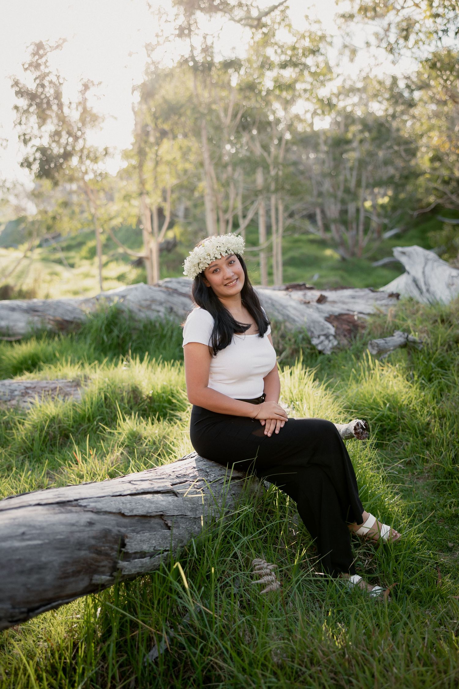 Young woman wearing flower crown sits on log in lush green park setting, smiling in white top and black pants.