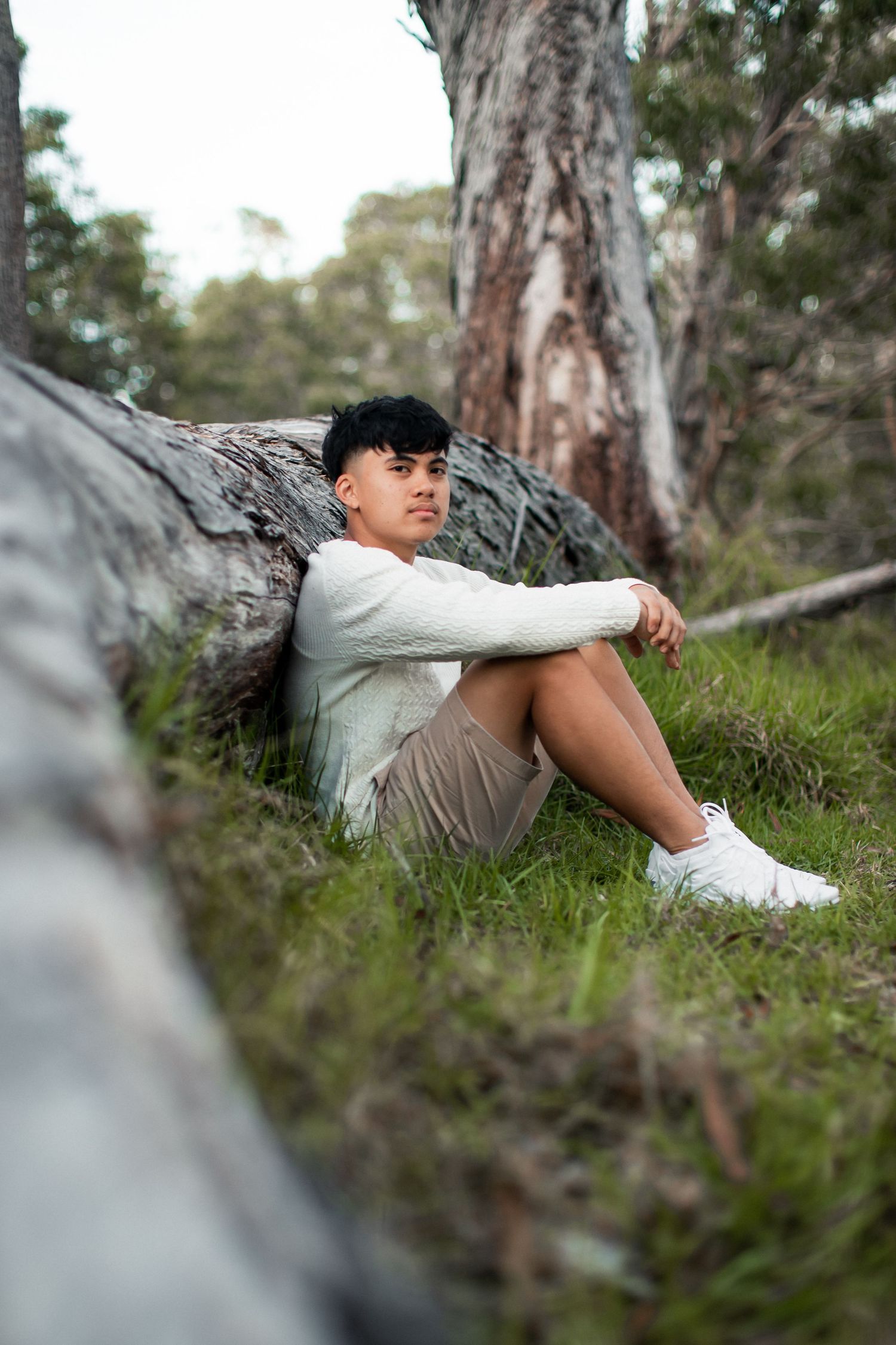 Young man sitting against a fallen log in a eucalyptus forest, wearing white sweater and khaki shorts.