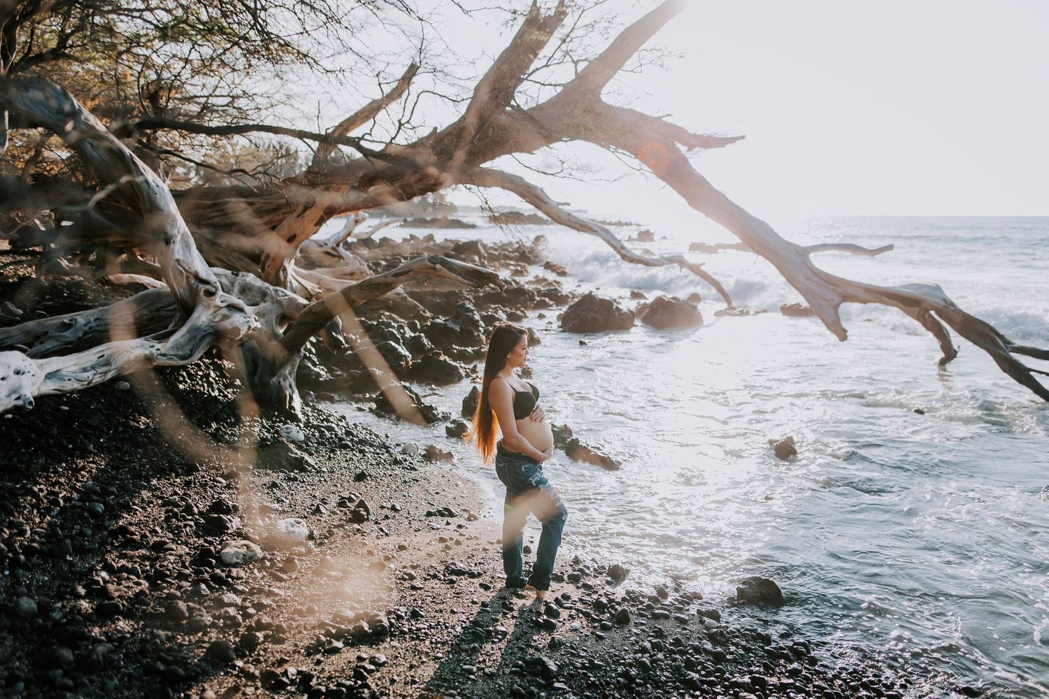 Woman standing on rocky shoreline beneath driftwood tree with sunlit ocean waves in background.