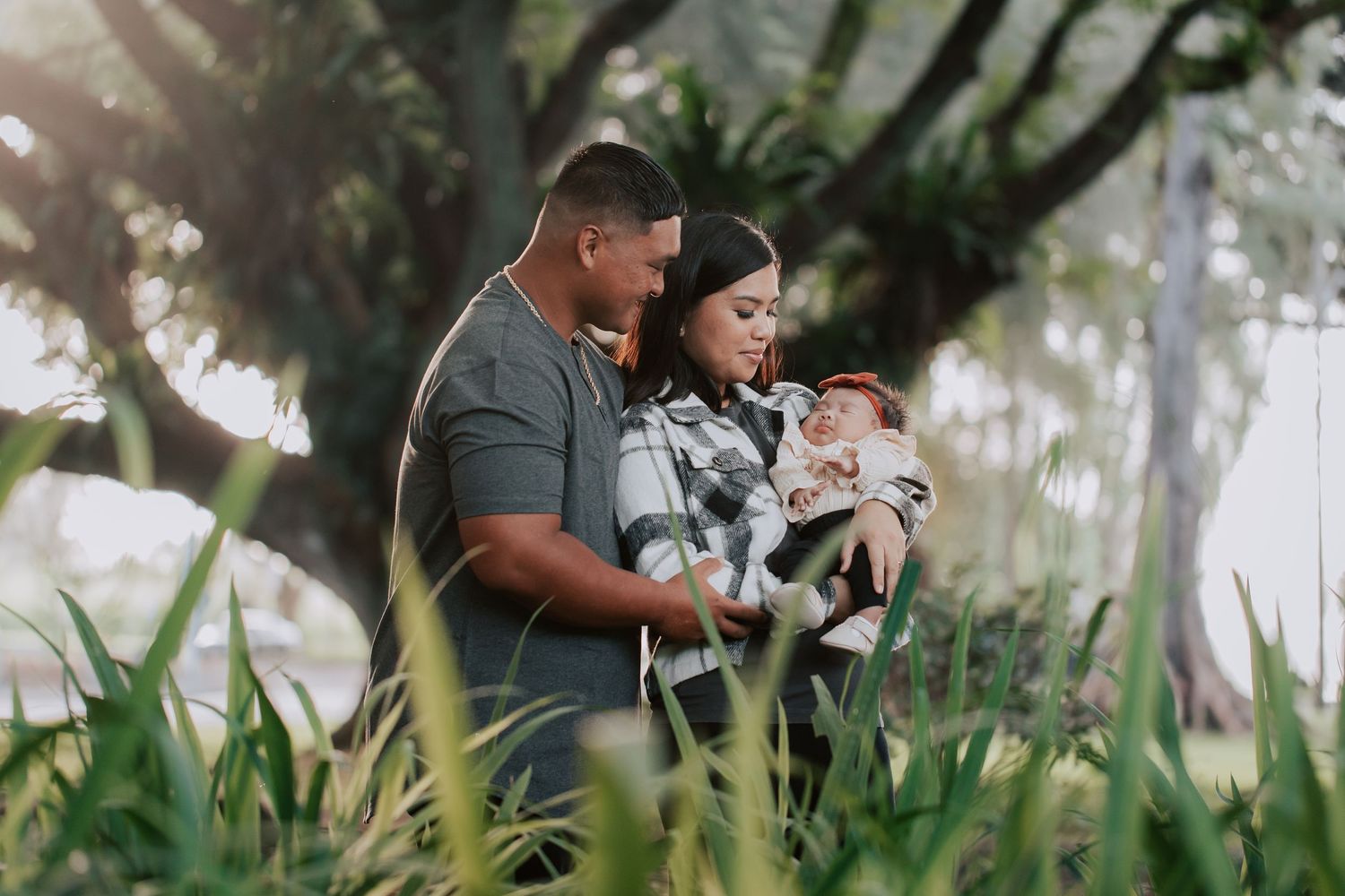 A couple stands outdoors holding their newborn baby surrounded by lush green tropical plants in soft natural light.