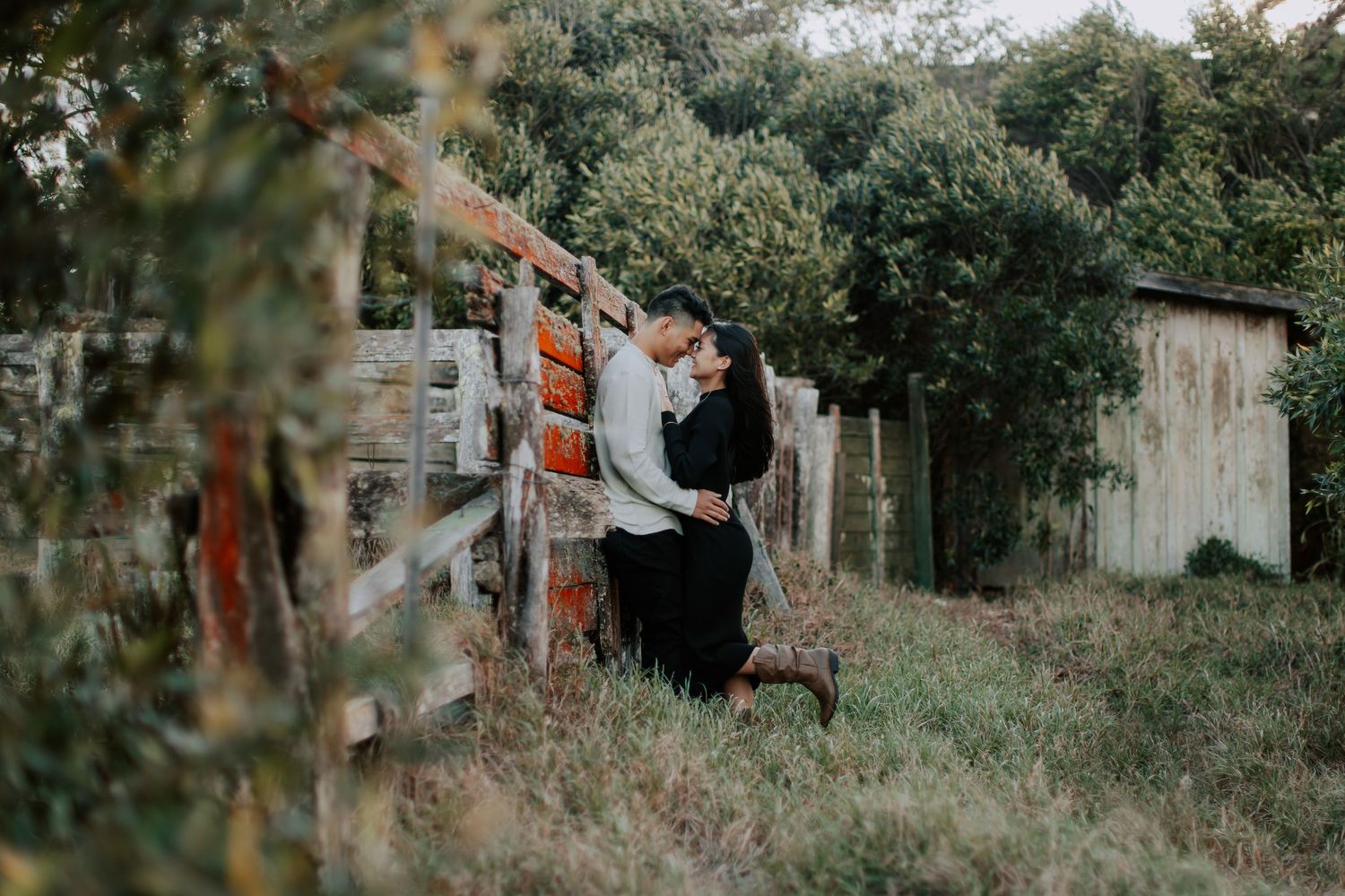 Couple sharing a romantic kiss near a rustic wooden fence surrounded by lush greenery and pumpkins outdoors.