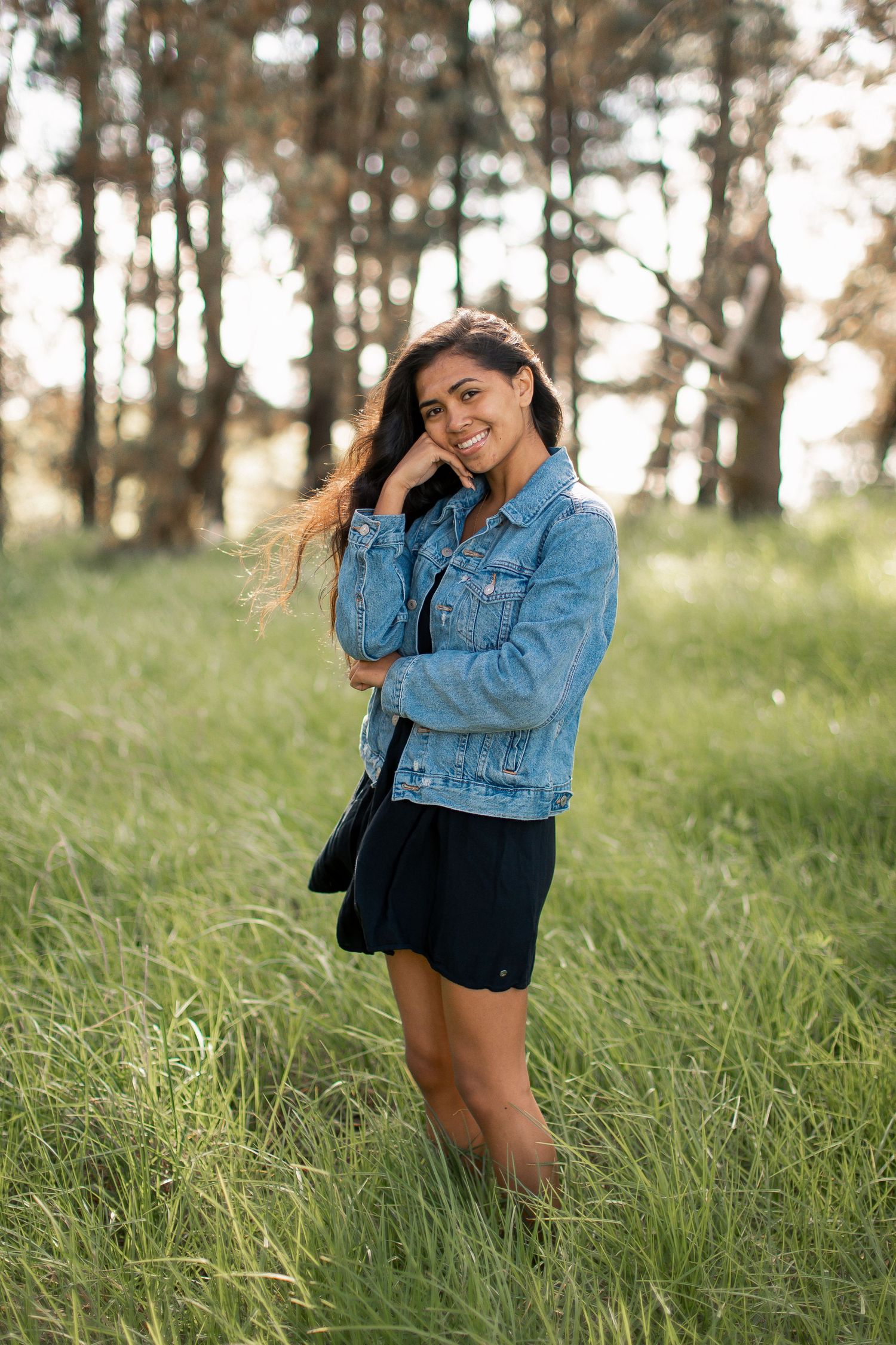 Young woman in denim jacket and black skirt smiling in a lush green meadow with tall trees in the background.