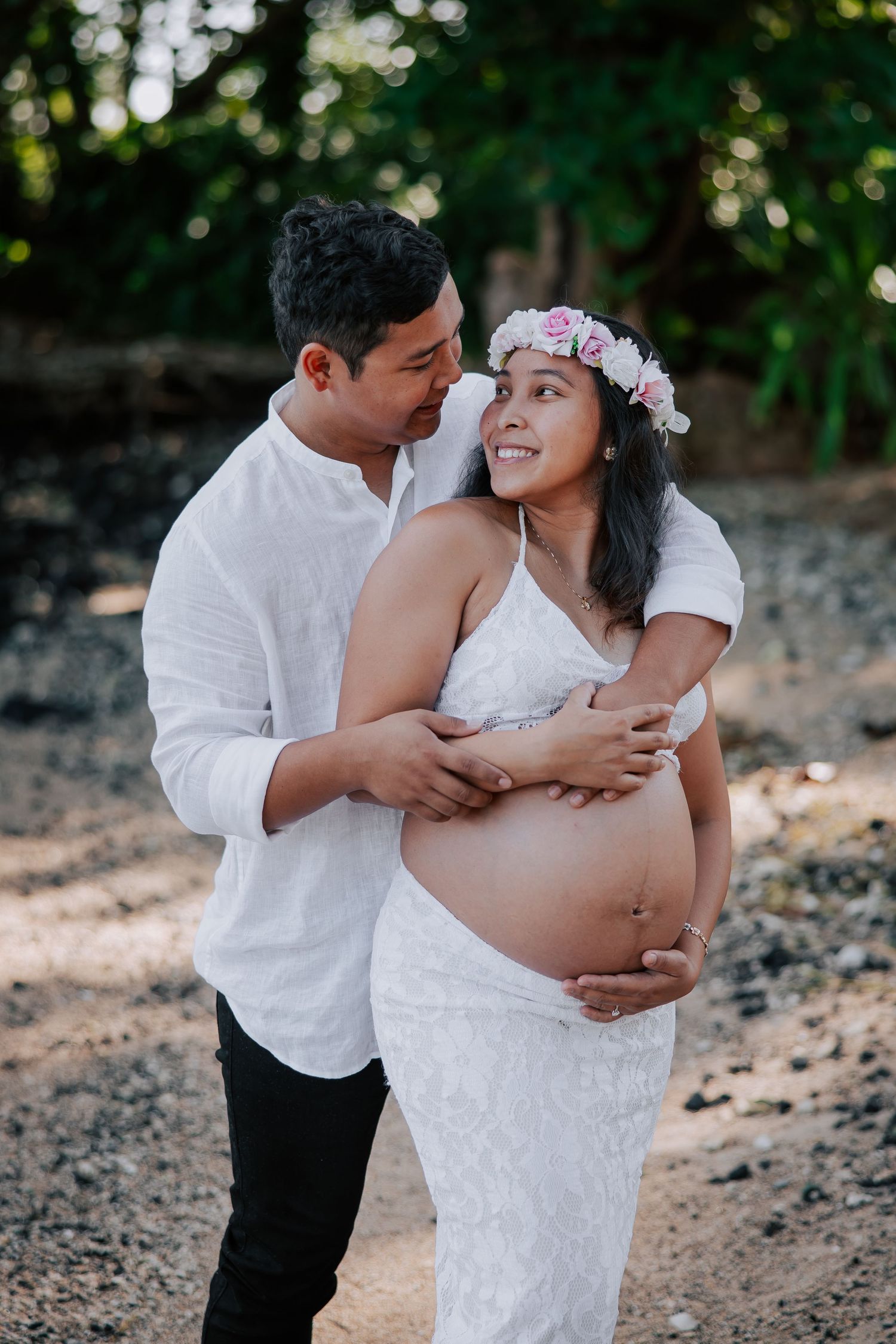 Couple in white maternity photoshoot outdoors, woman wearing floral crown with exposed baby bump, man kissing her cheek.
