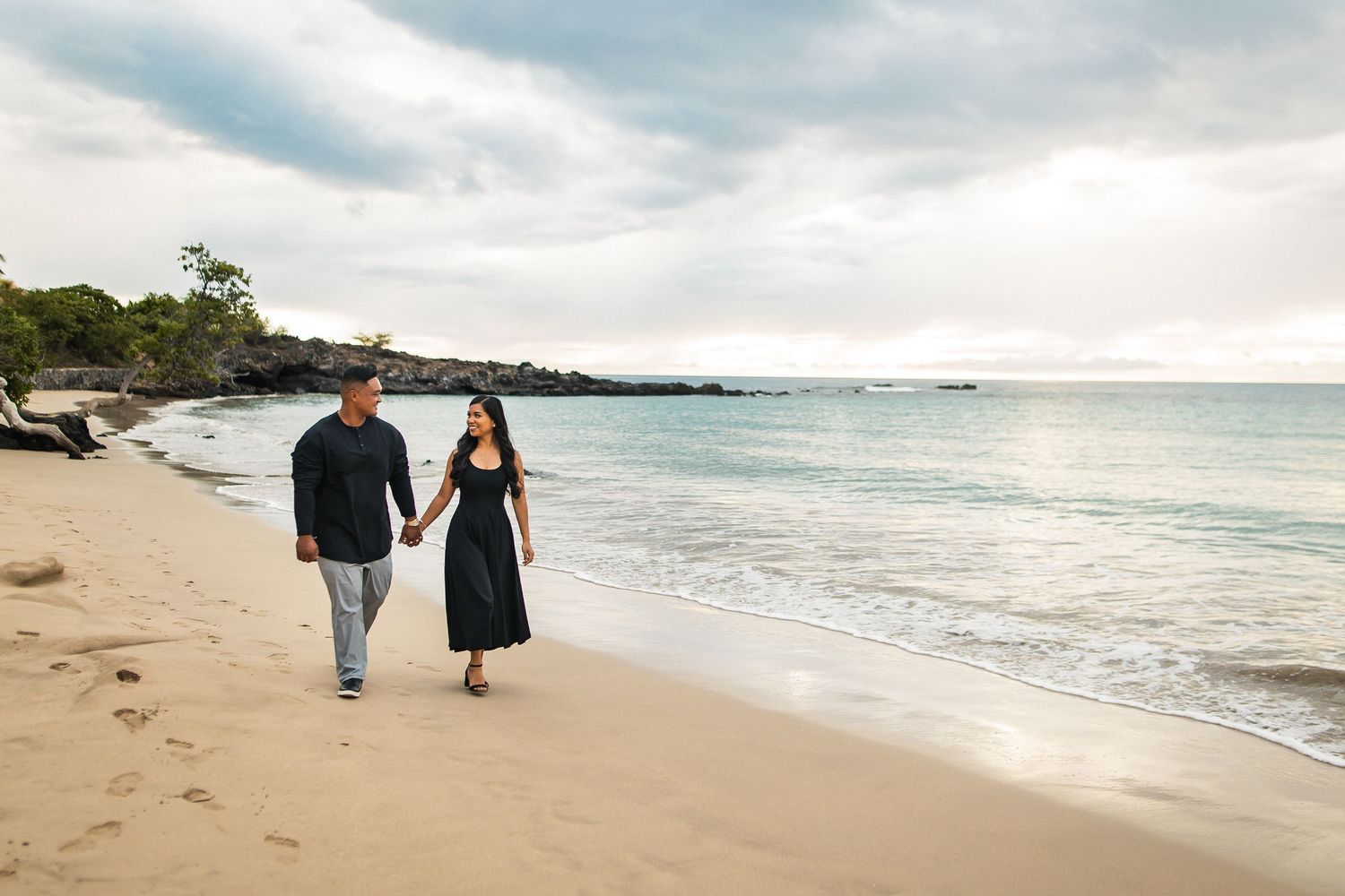 Couple holding hands walking on a sandy beach in matching black outfits with ocean waves and cloudy sky.