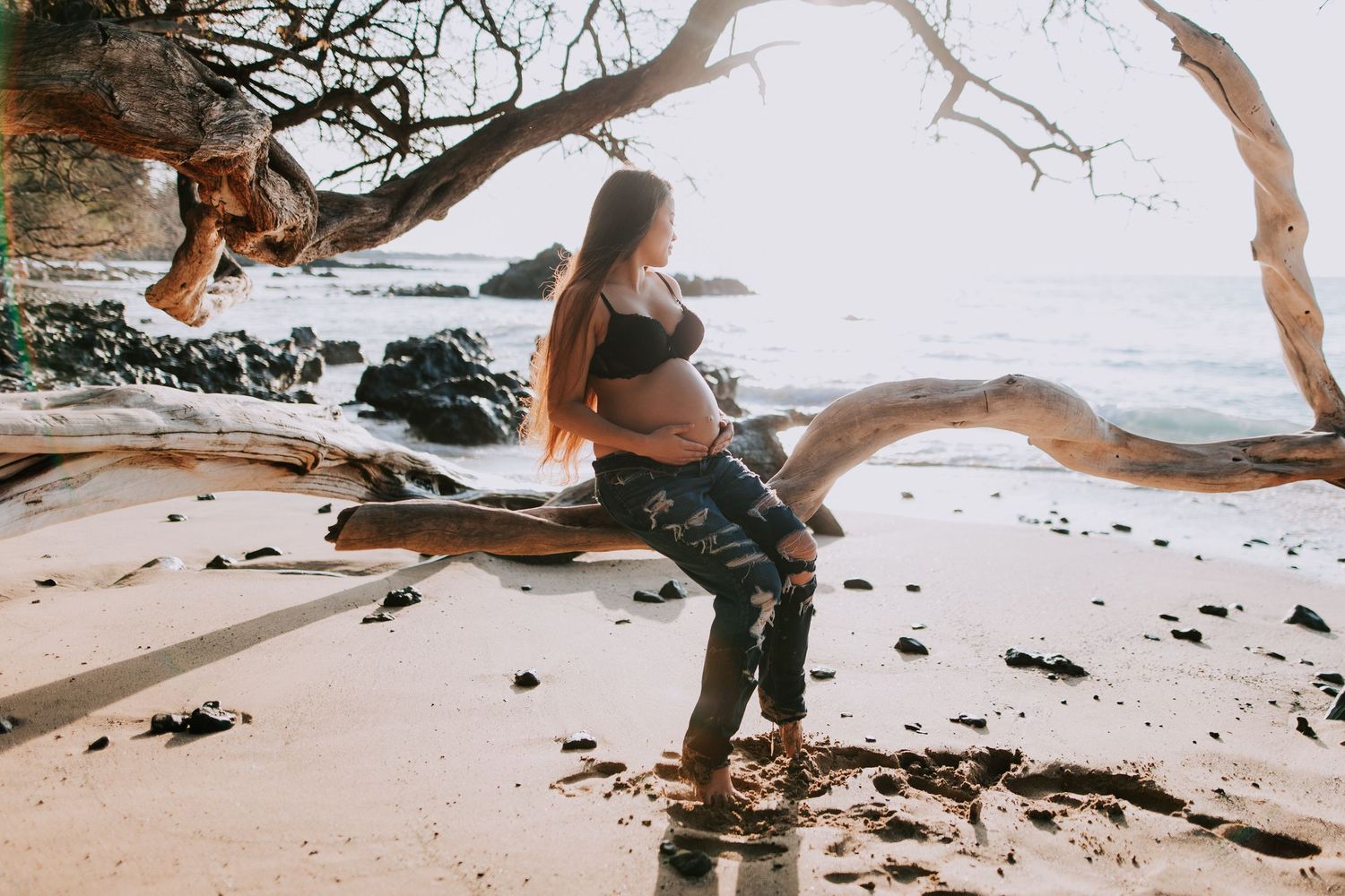 Pregnant woman in black bikini top sitting on driftwood at tropical beach during golden hour.
