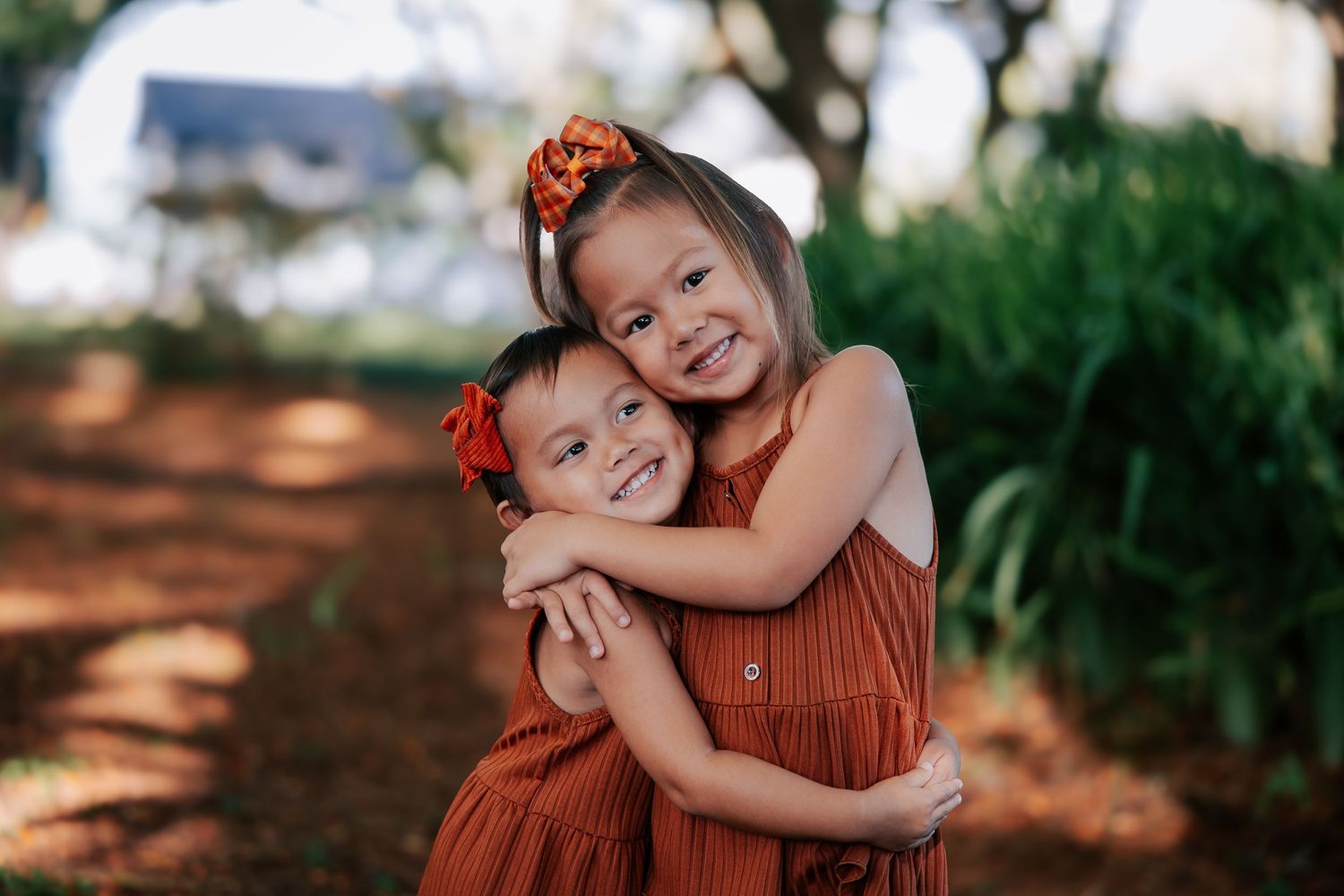 Two young sisters in matching rust-colored dresses share a warm hug outdoors, smiling joyfully with floral hair accessories.