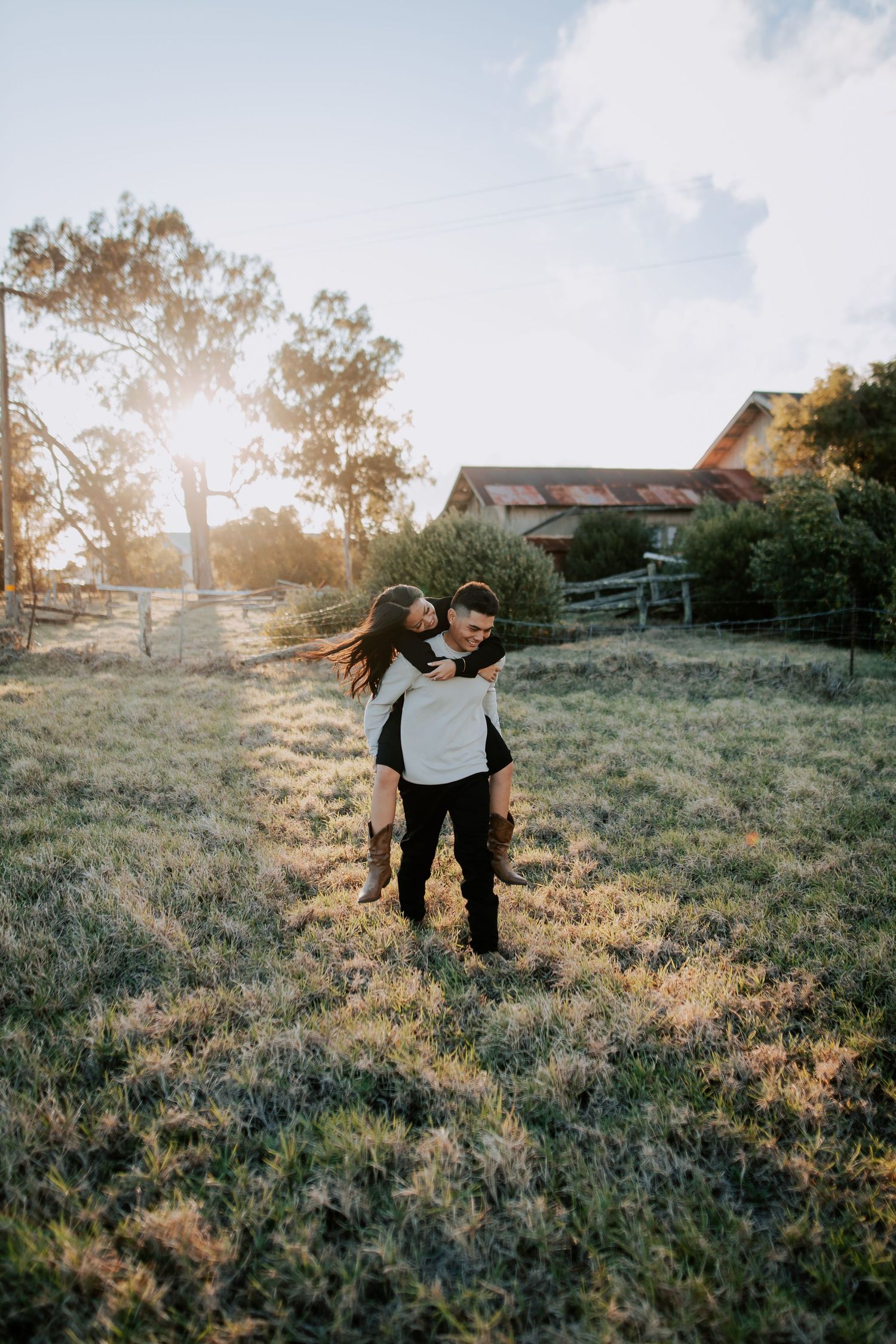 Couple sharing a piggyback ride in a sunlit field with a rustic barn and trees in the background.