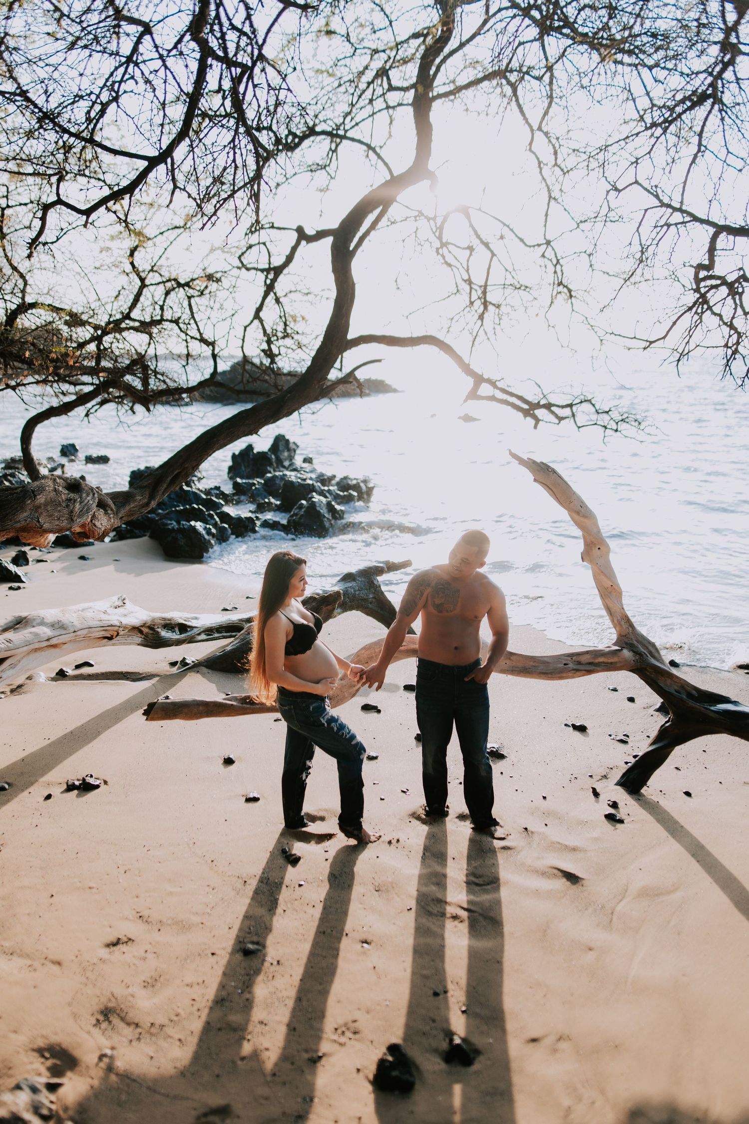 Couple holding hands on a sandy beach framed by bare tree branches with ocean waves and rocks in background.