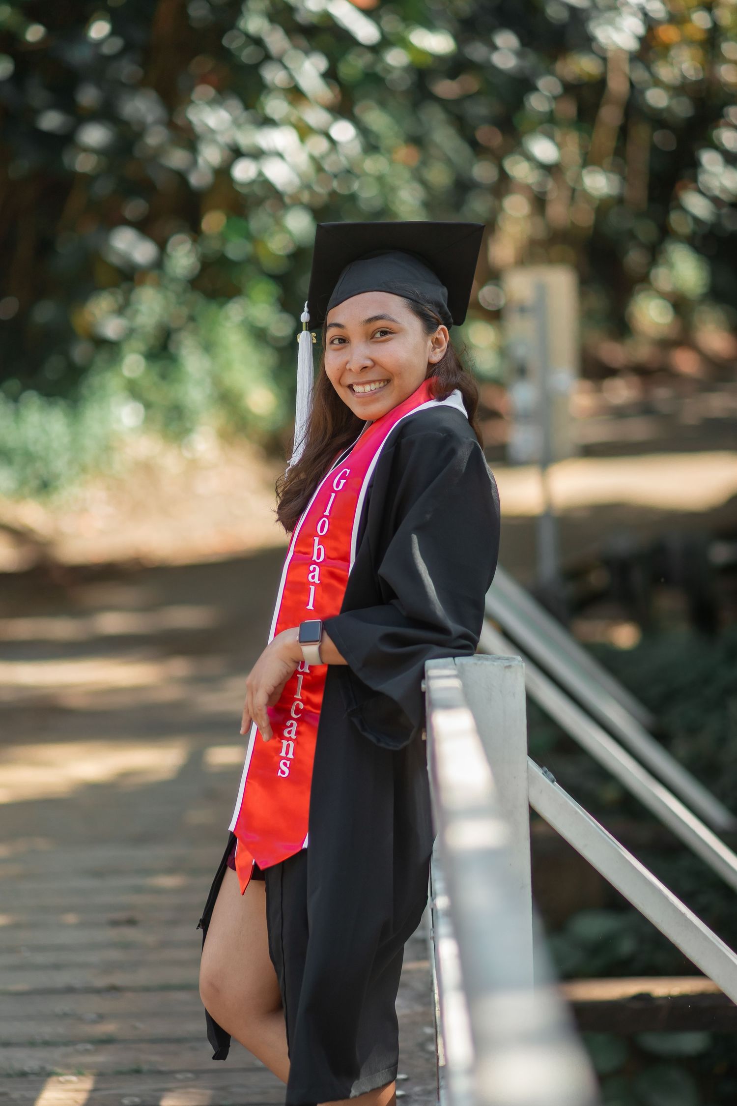 Smiling graduate in black cap and gown with red Global Class sash poses on a bridge surrounded by trees.